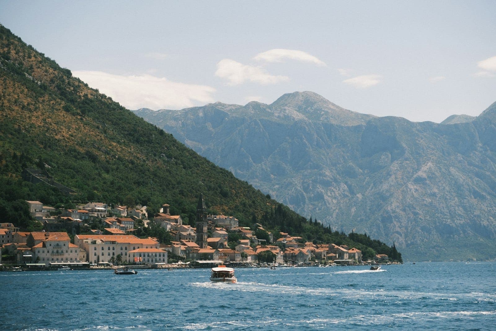 Vue depuis l'eau d'un bateau sur la Baie de Kotor, avec des bâtiments aux toits rouges et de hautes montagnes en arrière-plan.