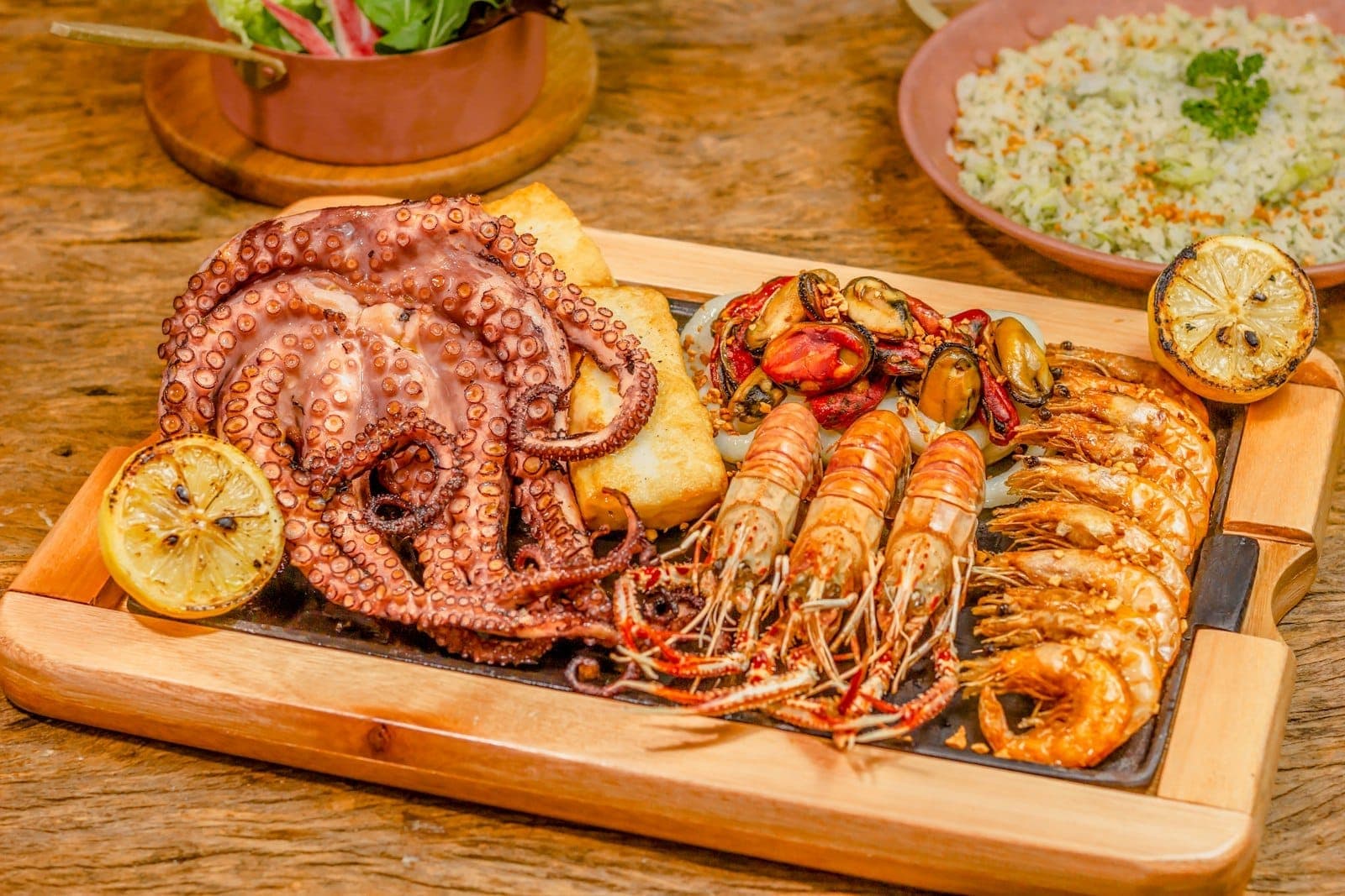 Una bandeja de mariscos frescos a la parrilla con pulpo, gambas, mariscos y limón, servida en una tabla de madera en un restaurante rústico.