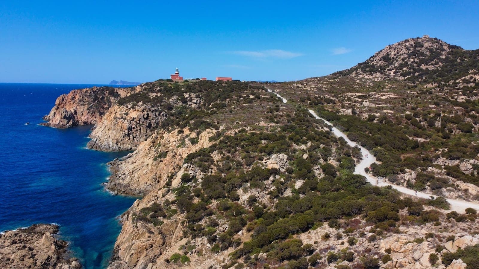 Paisaje costero rocoso con una carretera sinuosa y un faro a lo lejos en un día soleado en Cerdeña