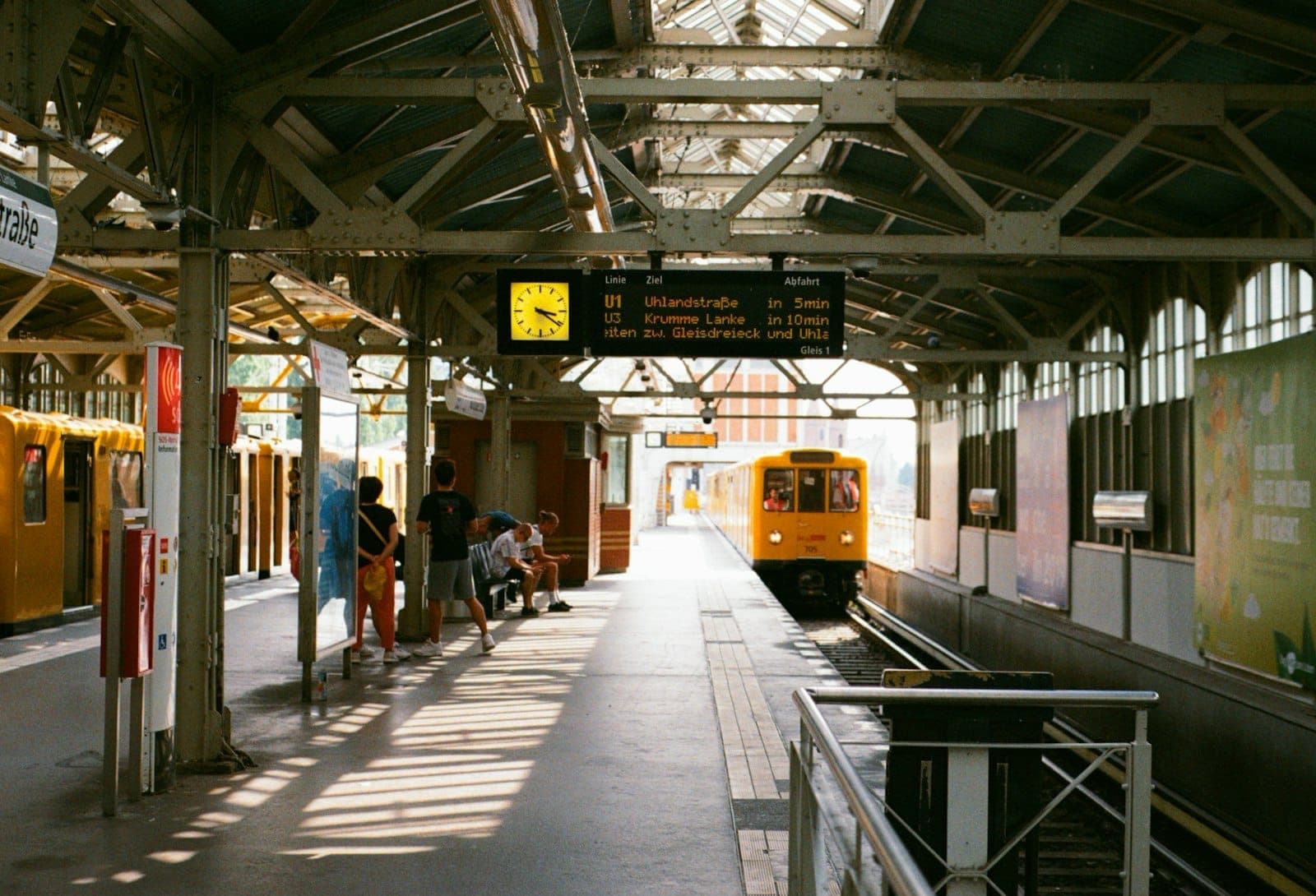 Stazione della U-Bahn di Berlino con treno giallo in arrivo, persone in attesa sulla banchina coperta e display degli orari in alto.