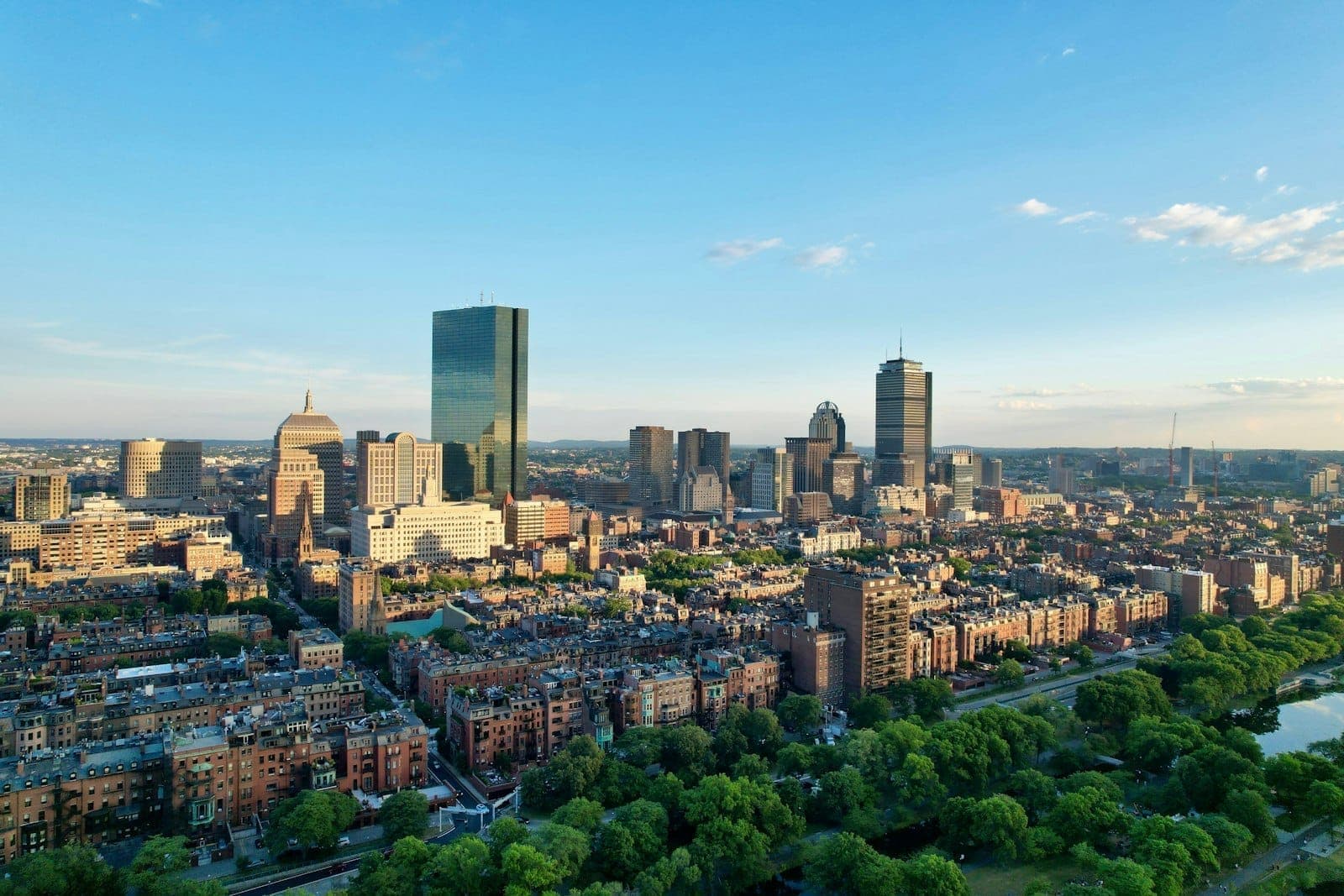 Vue aérienne du centre-ville de Boston avec des espaces verts, des bâtiments historiques en brique et des gratte-ciel modernes sous un ciel bleu dégagé.