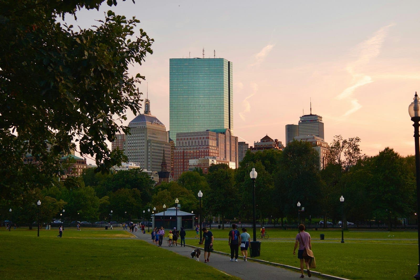 Boston Common au coucher du soleil avec la skyline en arrière-plan, des personnes se promenant sur les allées, des arbres verts et un espace de parc ouvert.