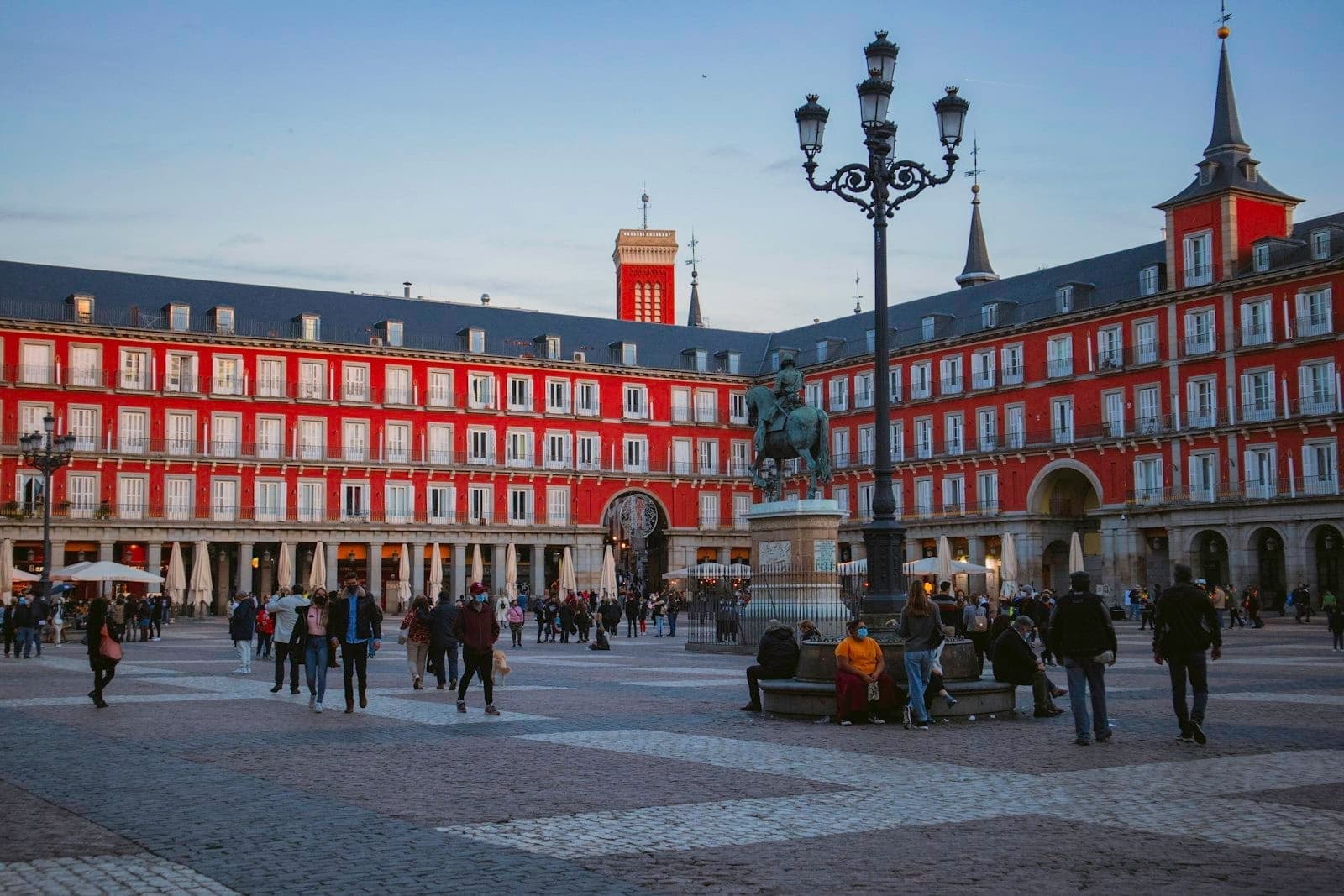 Vista serale della Plaza Mayor di Madrid con edifici rossi, statua equestre centrale, lampione e persone che passeggiano e siedono nella piazza.