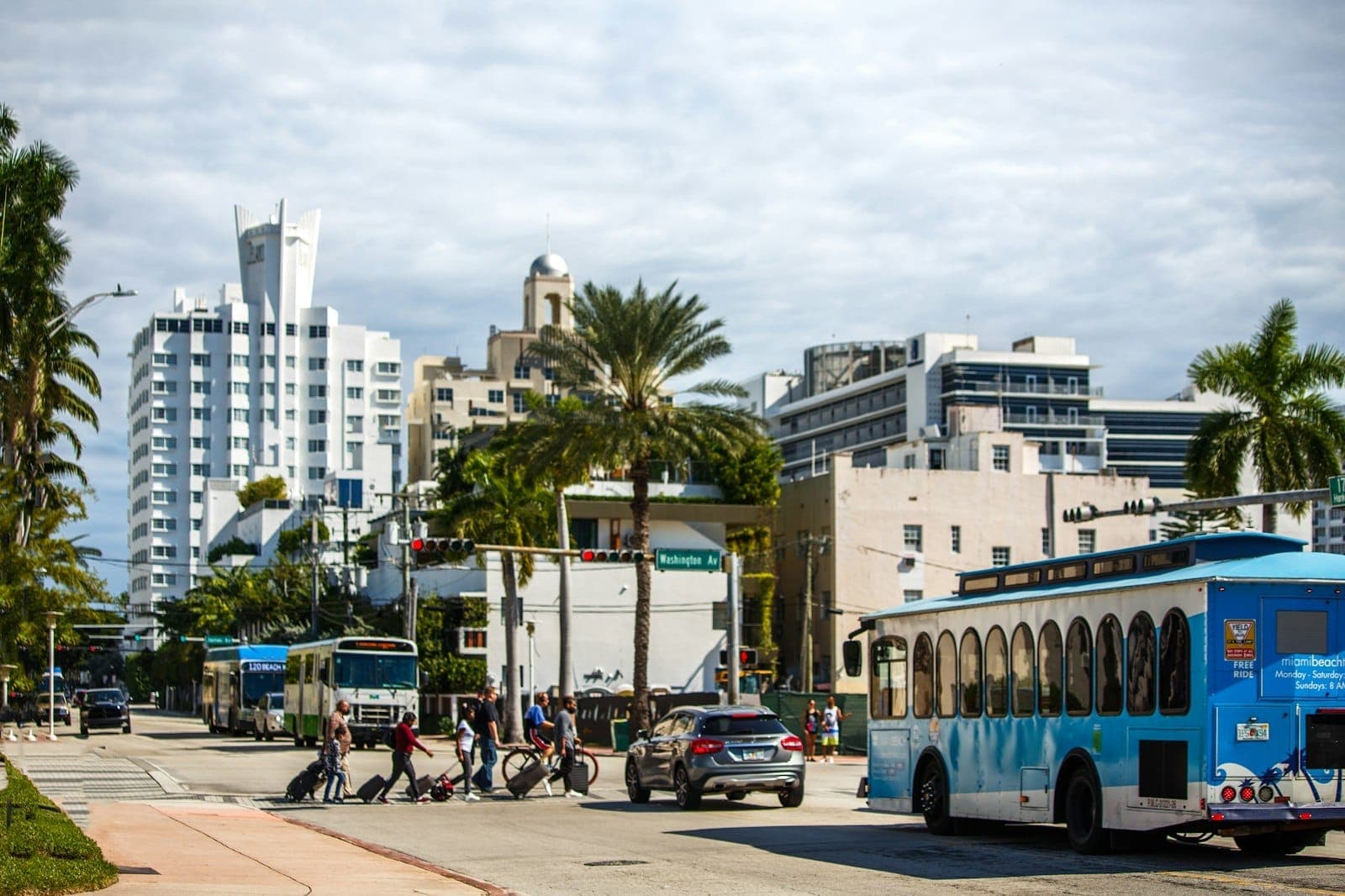 Rue à Miami avec des palmiers, un trolleybus de la ville, des piétons, des vélos et des bâtiments Art Déco sous un ciel partiellement nuageux.