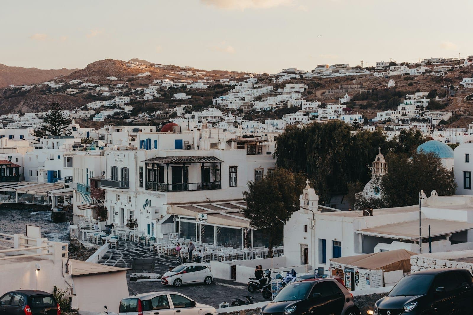 Blick auf die Stadt Mykonos mit geparkten Autos, weiß getünchten Gebäuden und Hügeln im Hintergrund bei Sonnenuntergang