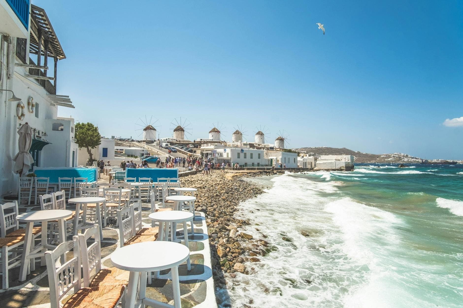 Küstenblick auf Mykonos Town mit ikonischen weißen Gebäuden, bunten Balkonen, Cafés im Freien und den berühmten Windmühlen im Hintergrund unter einem klaren blauen Himmel.