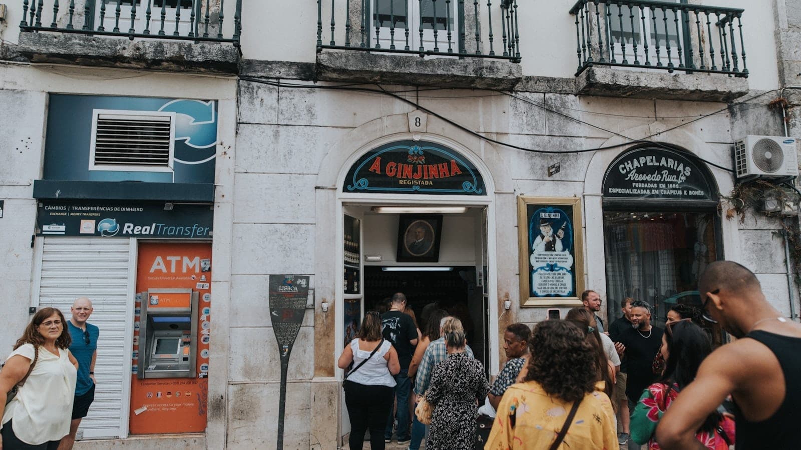Visitantes se reúnem do lado de fora da histórica entrada do bar A Ginjinha em Lisboa, com sua placa característica e ornamentada fachada de pedra visíveis.