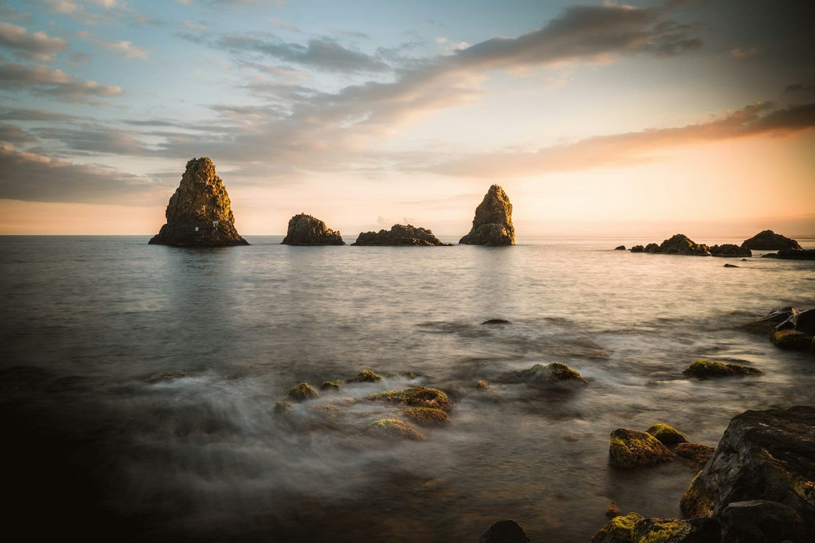Pemandangan tepi laut yang dramatis dengan formasi batuan basal gelap Kepulauan Cyclopean menjulang dari perairan Laut Ionia yang tenang saat matahari terbenam, cahaya oranye lembut menerangi pantai berbatu.