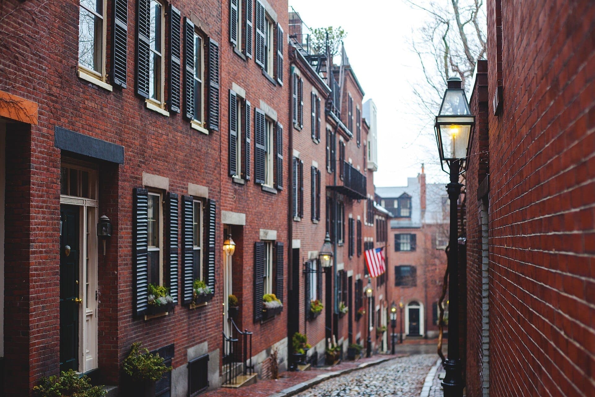 Des maisons en rangée en brique rouge avec des volets noirs longent les pavés d'Acorn Street à Boston, avec des lampadaires à gaz brillant sous une lumière diurne couverte.