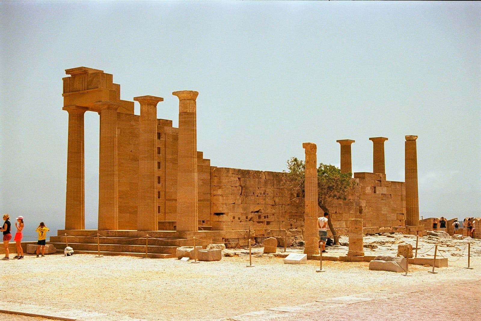 Ancient stone columns and partial ruins of the Acropolis of Lindos under a sunny, clear sky, with tourists exploring the historic site.