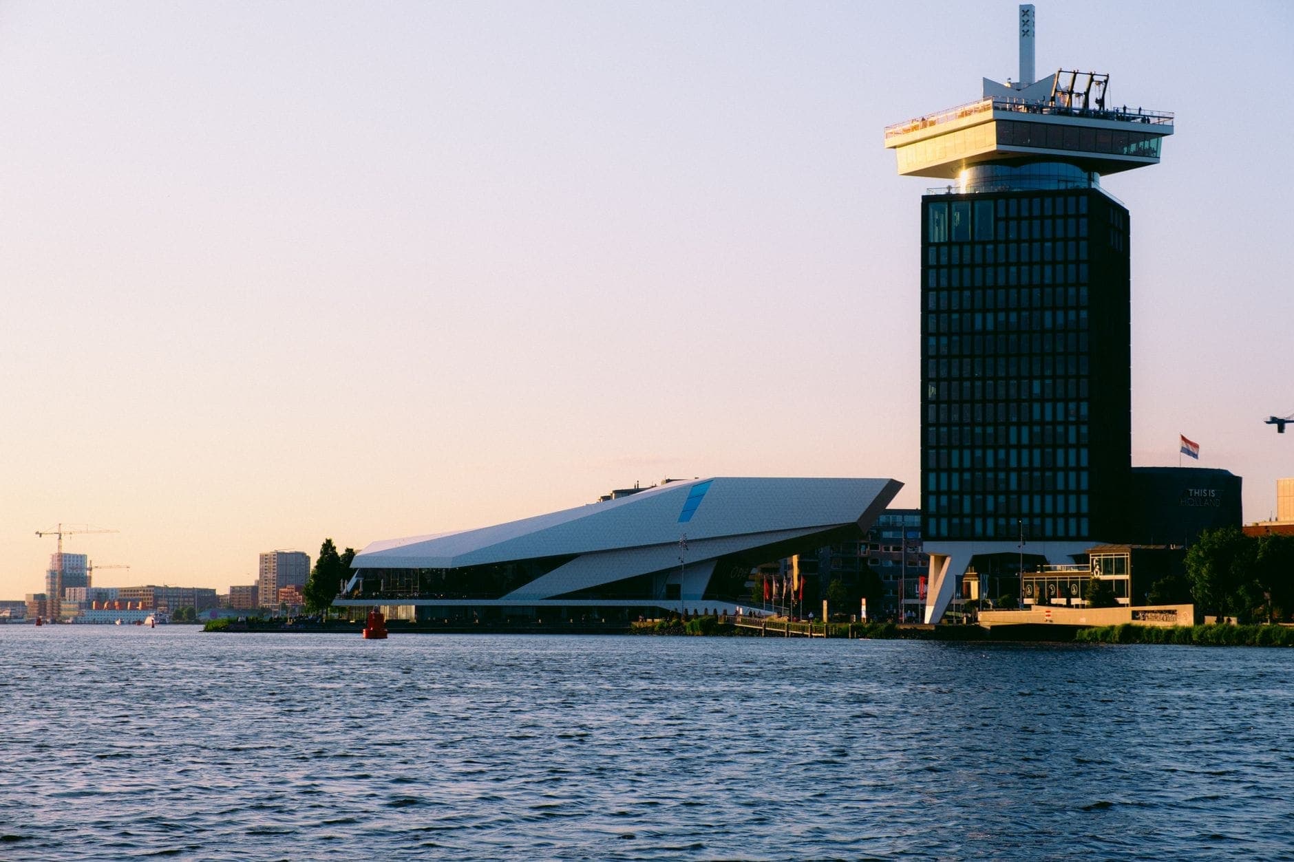 Der A'DAM Lookout Tower und das EYE Filmmuseum bei Sonnenuntergang, vom anderen Ufer des IJ aus gesehen, mit ruhigem Wasser und warmem Licht.