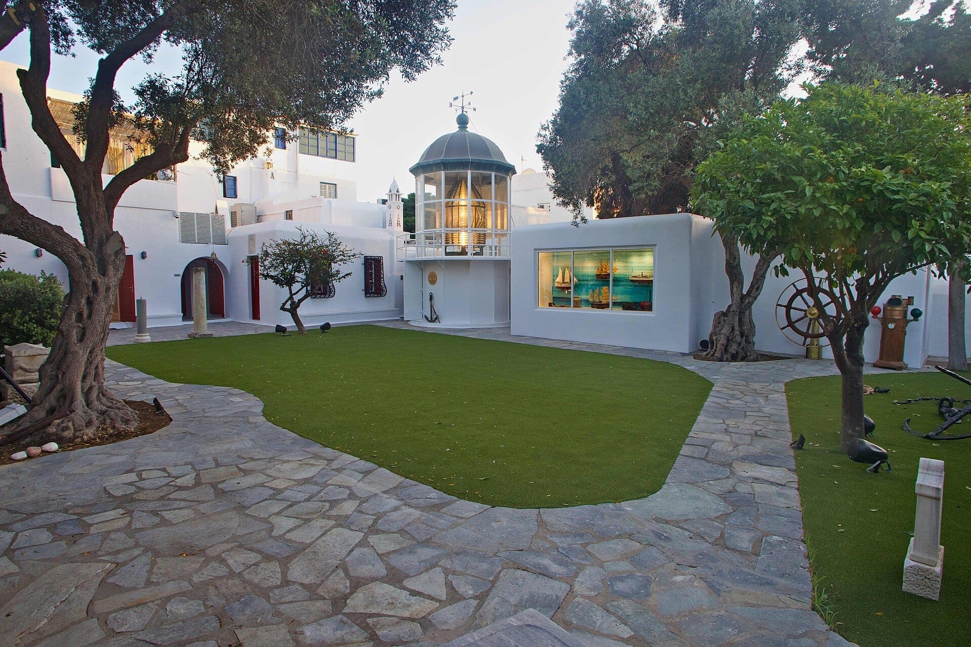Innenhof des Ägäischen Schifffahrtsmuseums in Mykonos mit weißen kykladischen Gebäuden, grünem Rasen, Bäumen und einem Fenster mit Schiffsmodellen.
