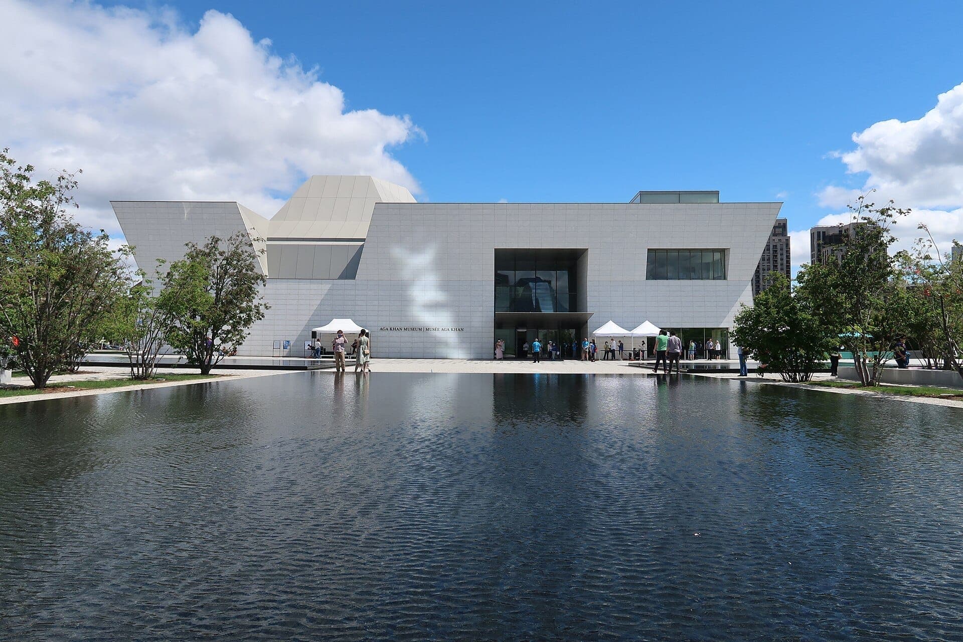 Moderno edificio bianco dell'Aga Khan Museum a Toronto, visto attraverso una vasca riflettente con visitatori all'esterno sotto un cielo azzurro brillante.