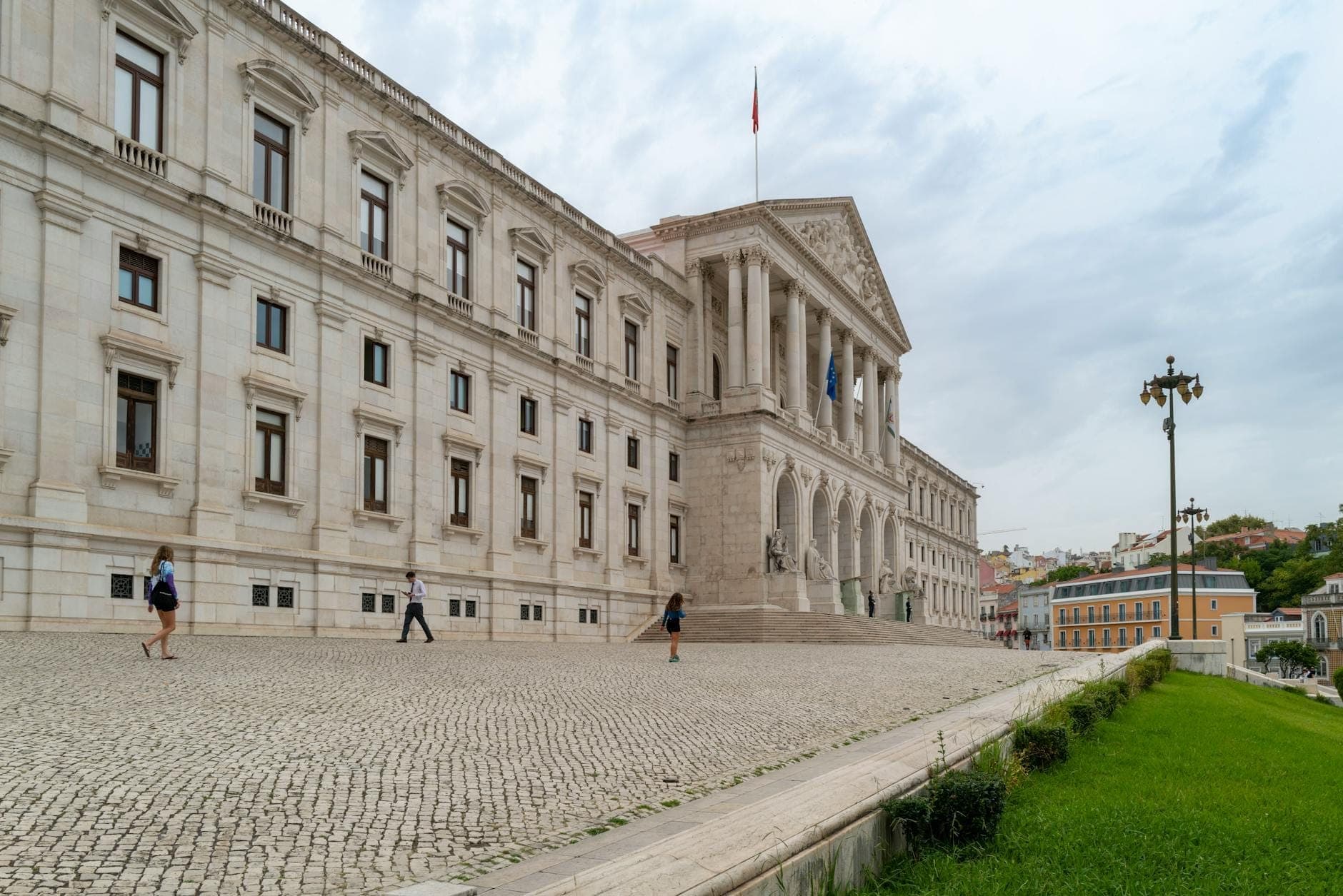 Vista ampla da fachada neoclássica do Palácio Nacional da Ajuda em Lisboa, com pedestres na praça de calçada portuguesa e gramado verde em primeiro plano sob um céu nublado.