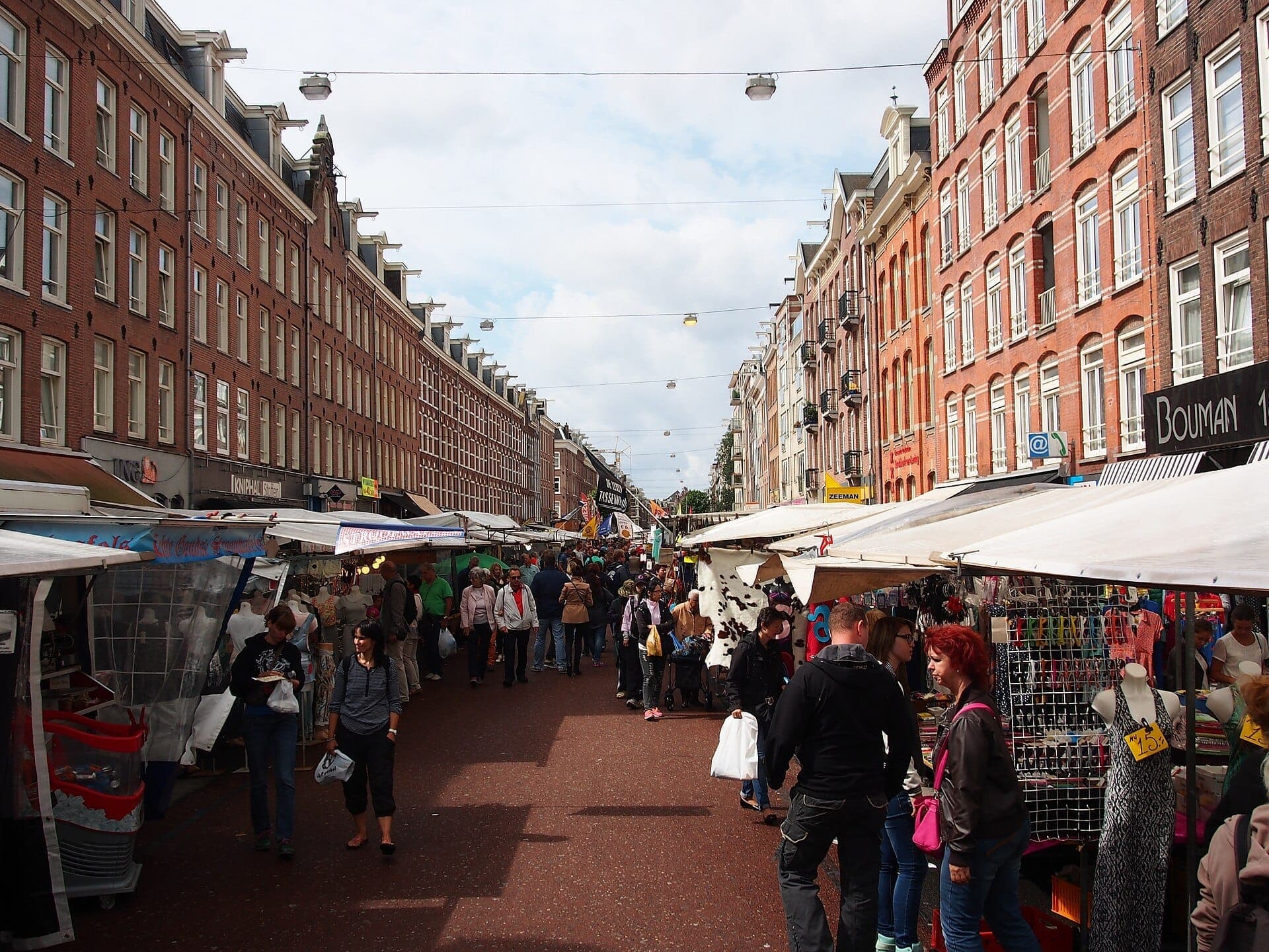 Besucher stöbern an bunten Ständen und Waren auf dem Albert Cuyp Markt in Amsterdam, gesäumt von historischen Backsteingebäuden unter einem strahlend blauen Himmel.
