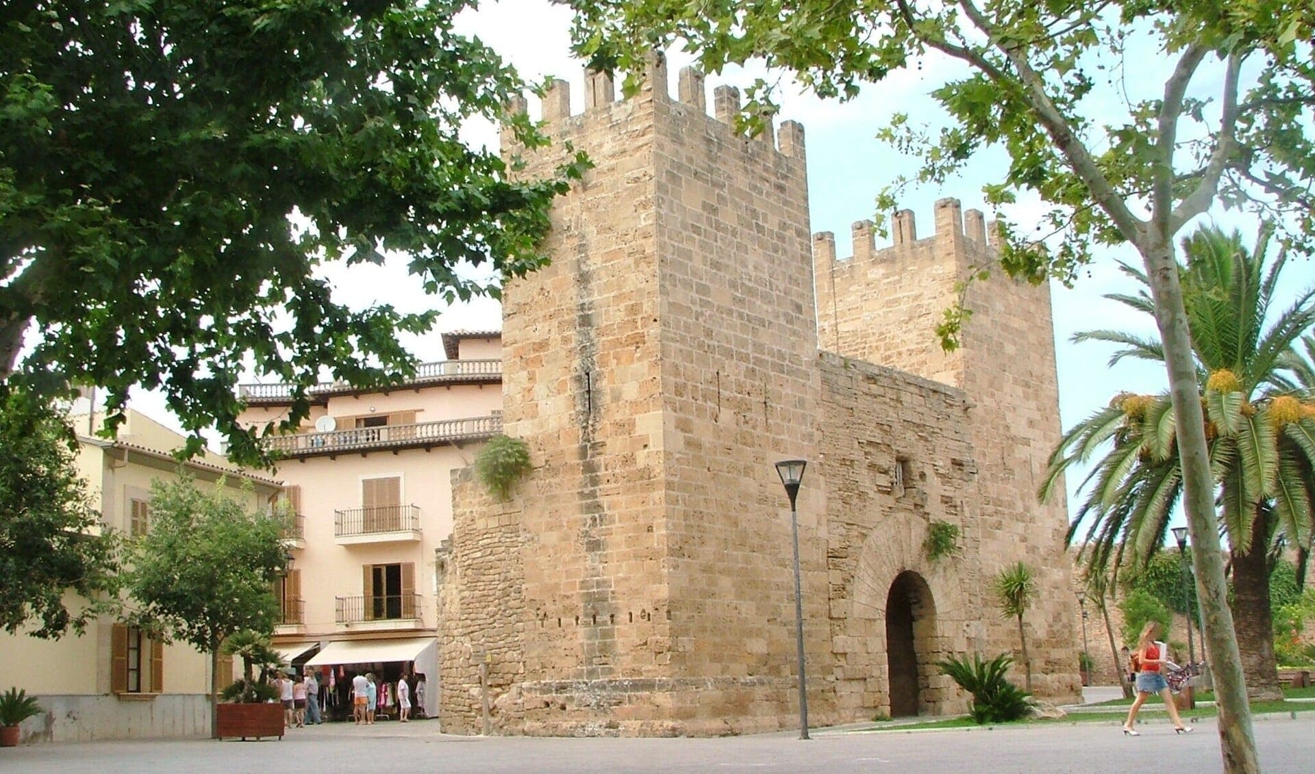 Steinerne mittelalterliche Stadtmauern und Türme der Altstadt von Alcúdia, Mallorca, umgeben von belaubten Bäumen und nahegelegenen historischen Gebäuden bei Tag.
