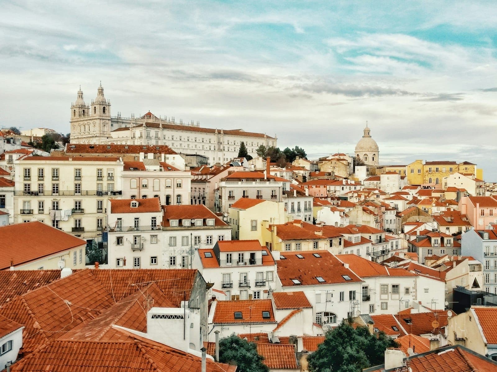 Vista panorâmica sobre os telhados vermelhos de Alfama, igrejas históricas e edifícios azulejados a estender-se até ao Rio Tejo, sob um céu luminoso em Lisboa, Portugal.