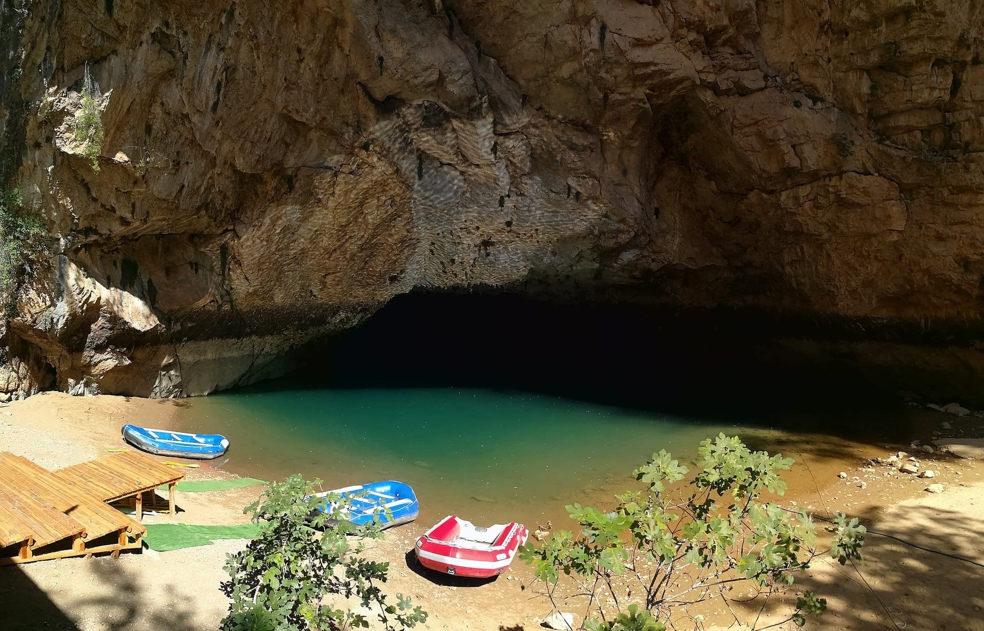 Eingang zur Altınbeşik-Höhle mit türkisfarbenem Untergrundse, rauen Steinwänden und mehreren bunten Schlauchbooten am sandigen Ufer.