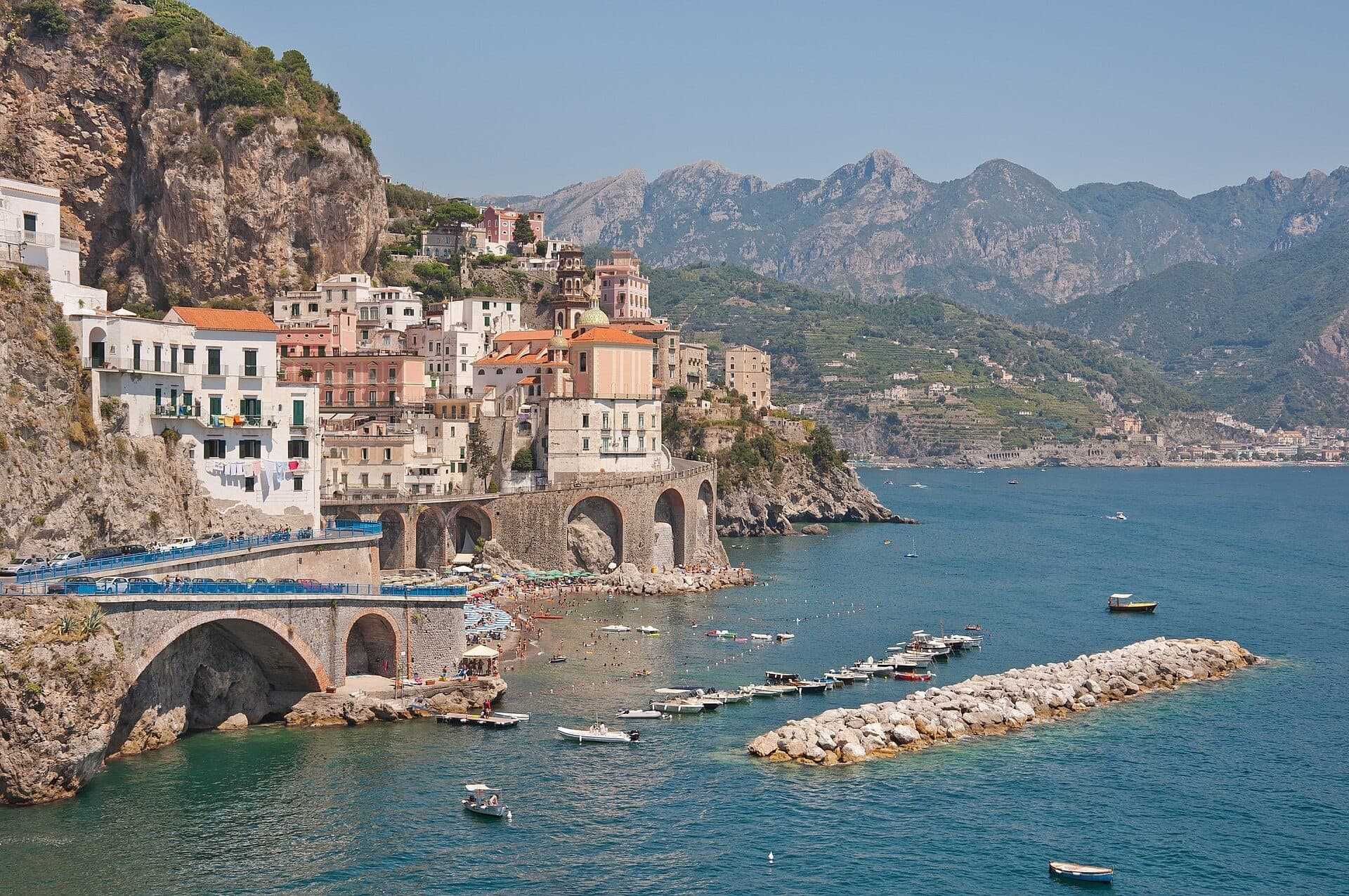 Casas coloridas de Atrani, na Costa Amalfitana, com vista para o mar azul, barcos na água e montanhas ao fundo.