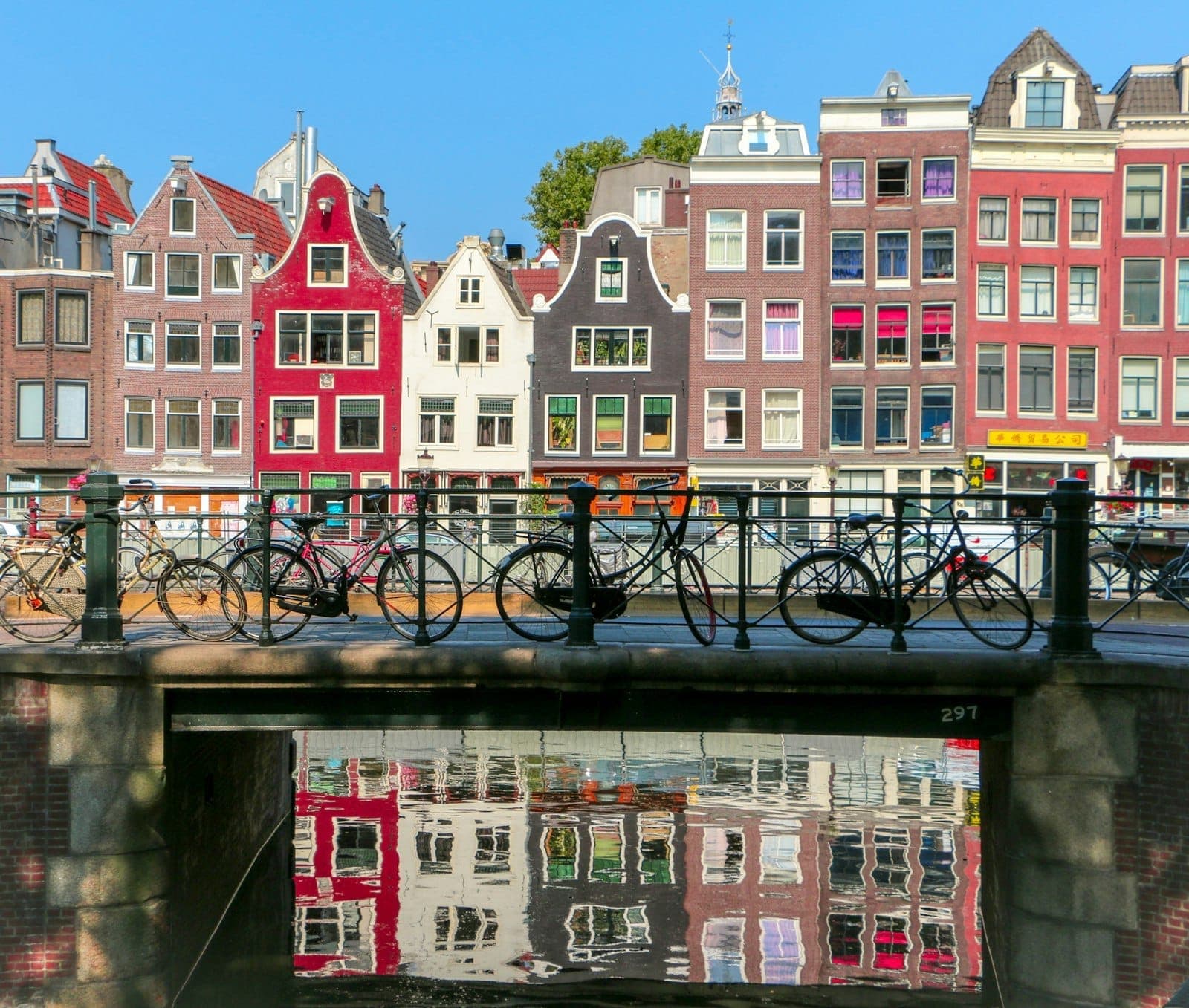 Bunte Grachtenhäuser mit traditionellen Fassaden, die sich im Wasser spiegeln, Fahrräder auf einer Brücke in Amsterdam an einem sonnigen Tag.
