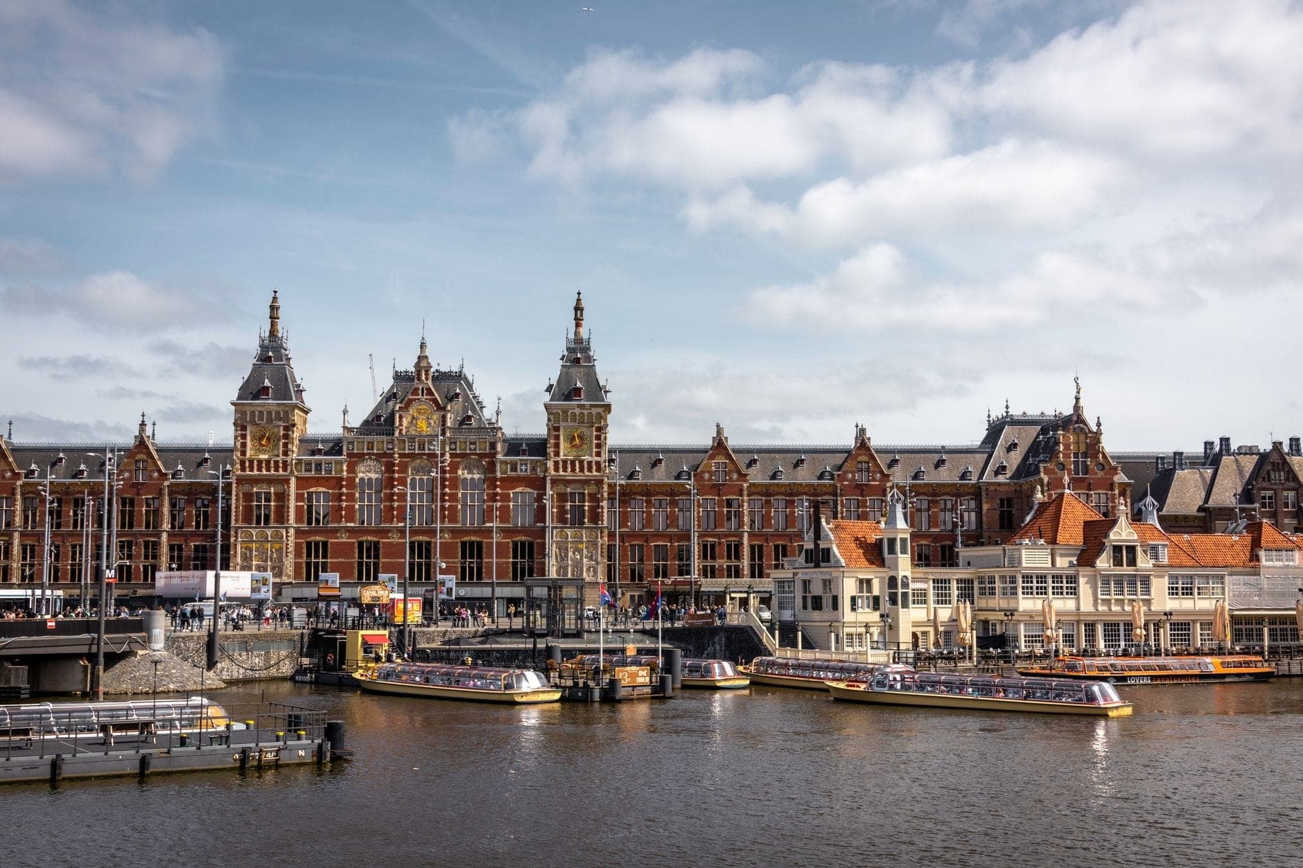 Amsterdam Centraal Bahnhof mit Kanalkreuzfahrtbooten, die im Vordergrund an den Anlegestellen festgemacht haben, unter einem teilweise bewölkten Himmel.