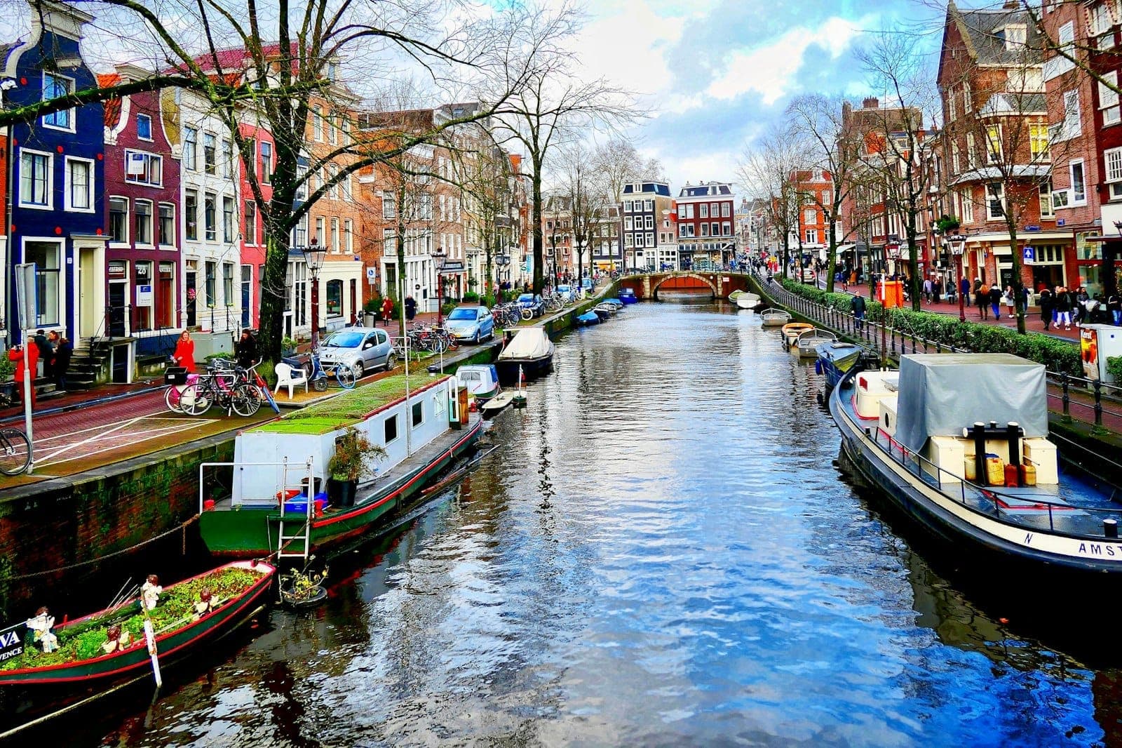 Helle Kanalszene in Amsterdam mit traditionellen schmalen Häusern, Booten und einem baumgesäumten Kanal unter blauem Himmel.