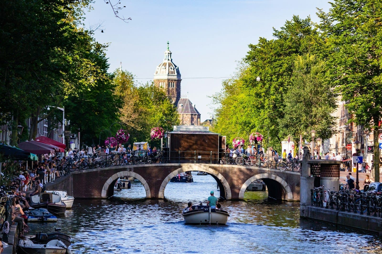 Belebter Amsterdamer Kanal mit Booten, einer mit Menschen überfüllten Steinbrücke, belaubten Bäumen und historischen Gebäuden im Sonnenlicht des Spätfrühlings oder Sommers.