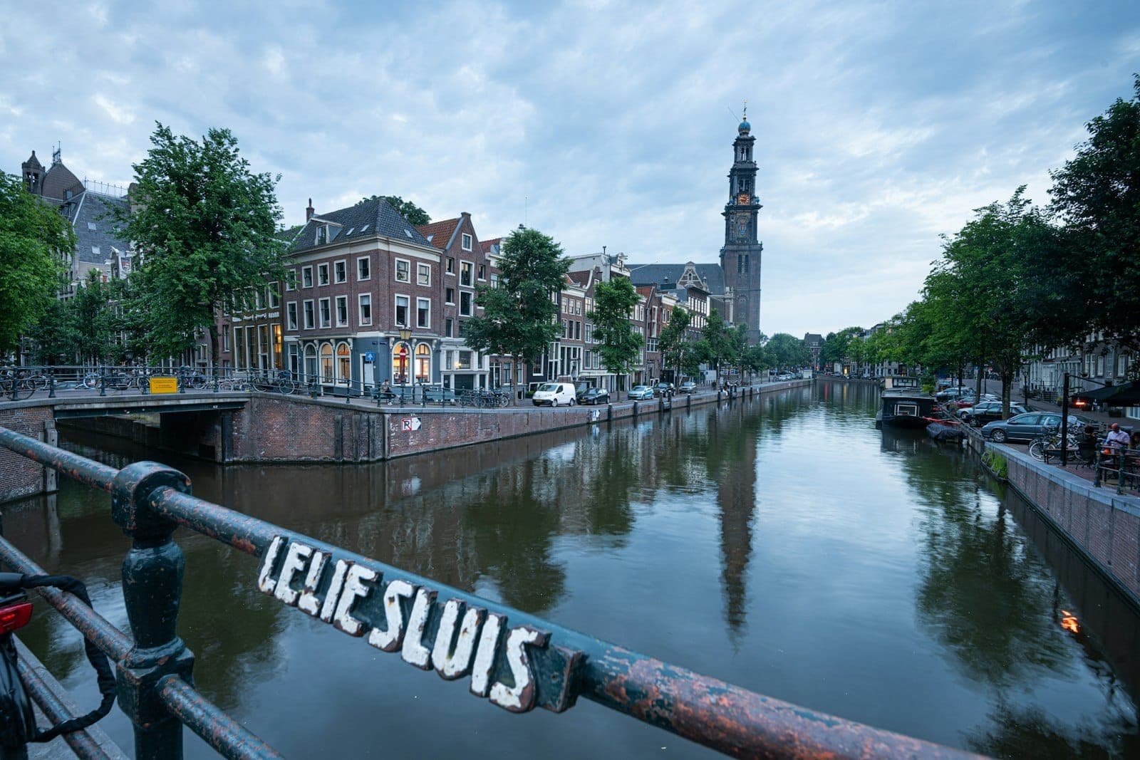 Klassischer Blick auf eine Amsterdamer Gracht mit historischen Häusern, Bäumen am Kanalufer und dem Kirchturm der Westerkerk, gesehen von einer Brücke mit einem Schild mit der Aufschrift 'Lelie Sluis'.