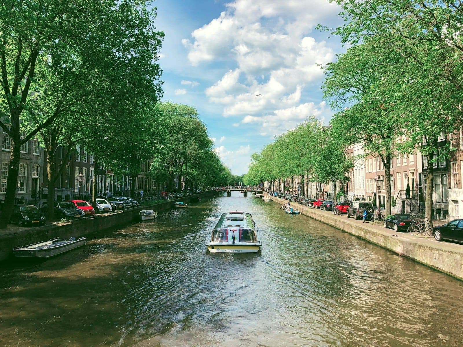 Ein malerischer Kanal in Amsterdam mit Bäumen auf beiden Seiten, am Wasser geparkten Autos und einem Boot, das die Mitte entlangfährt.