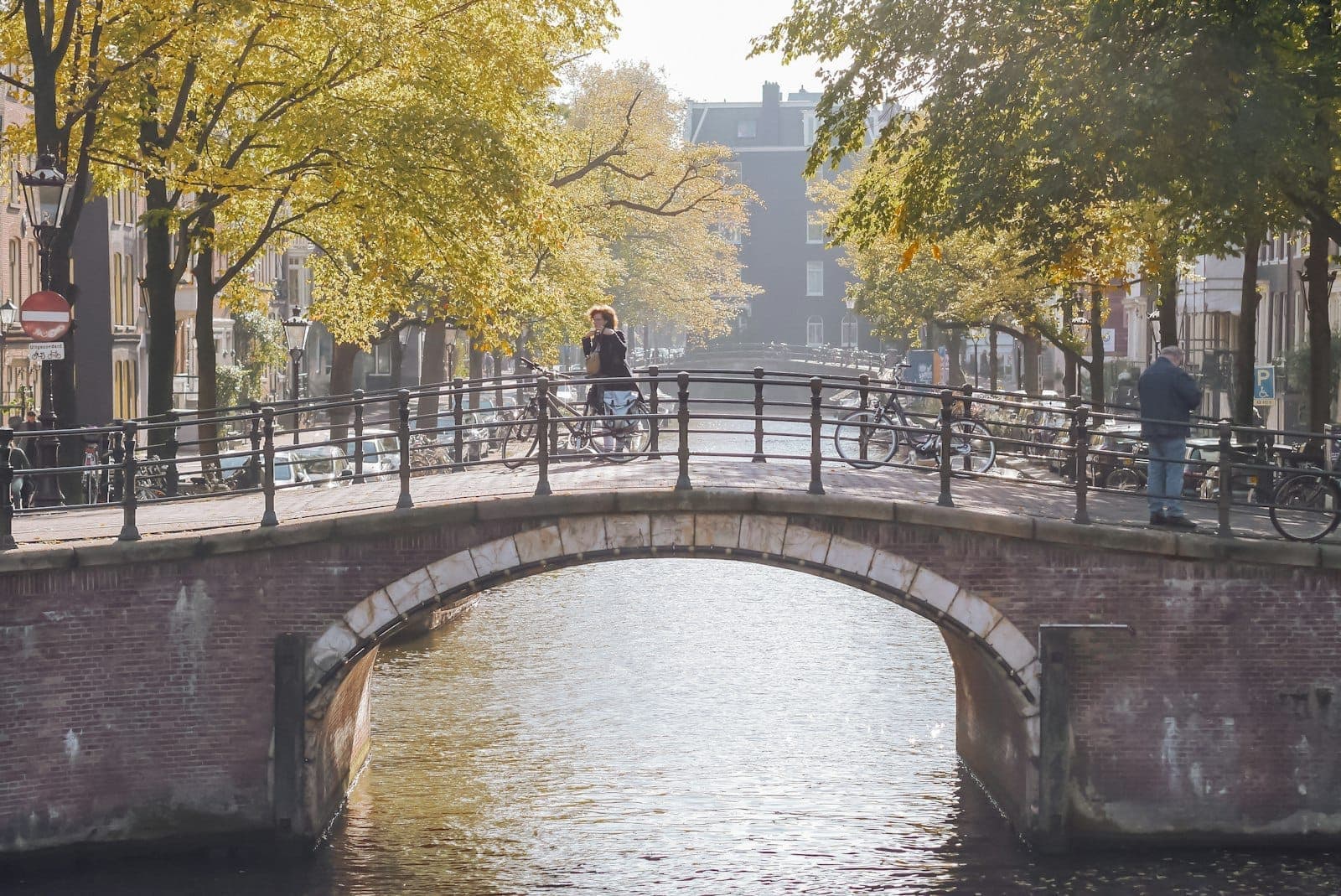 Sonnenbeschienene Kanalszene in Amsterdam mit einer Steinbrücke, spazierenden und radfahrenden Menschen sowie Bäumen entlang des Wassers.