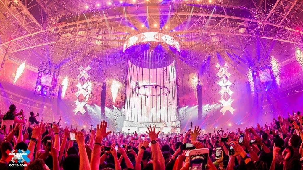 Eine große Menge tanzt unter lebhaften Bühnenlichtern und Amsterdam Dance Event Dekorationen in einem vollgepackten Musikclub während eines Live-Konzerts.