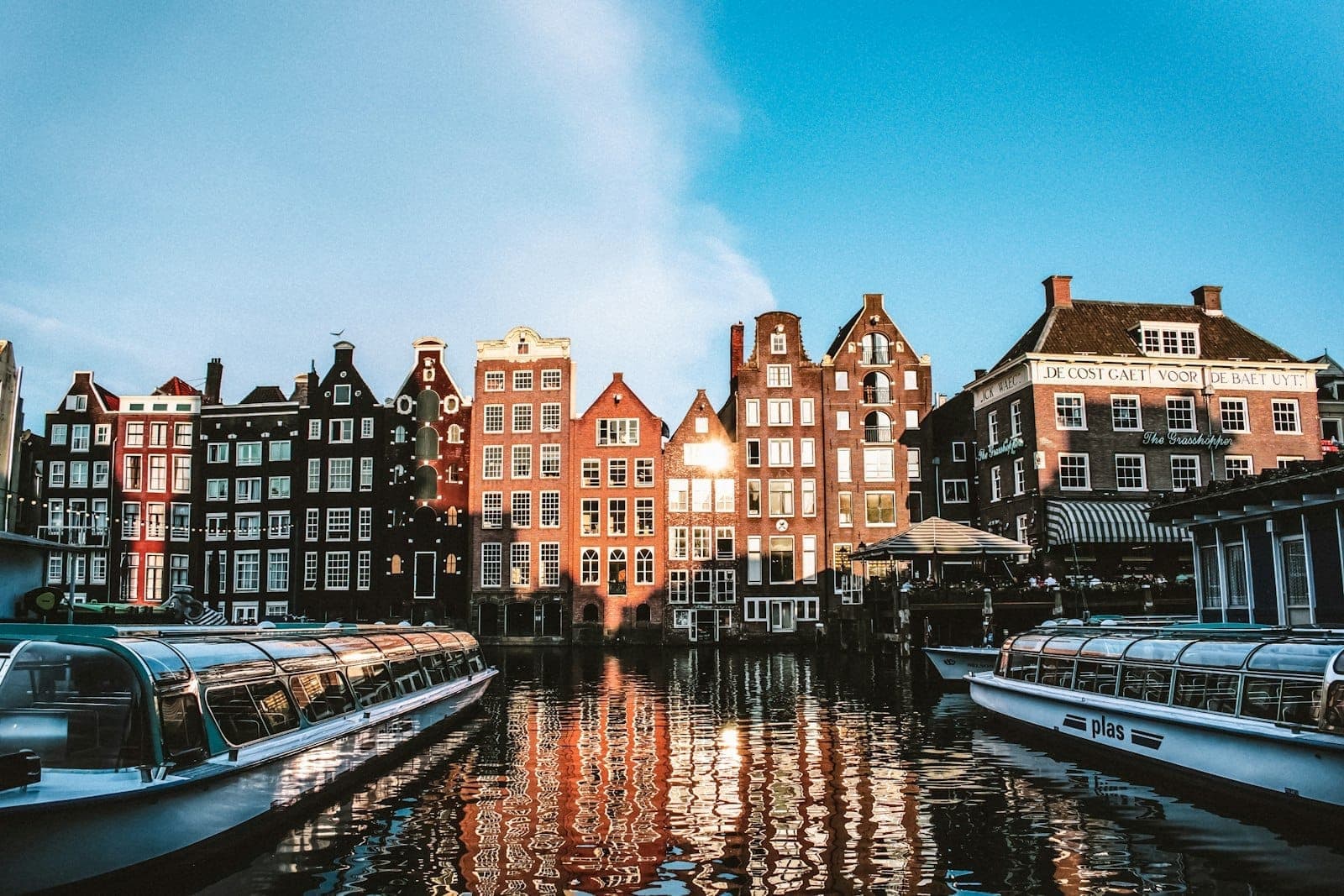 Klassische Kanalszene in Amsterdam mit traditionellen niederländischen Häusern und zwei Kanalkreuzfahrtbooten mit Glasdach bei Sonnenuntergang.