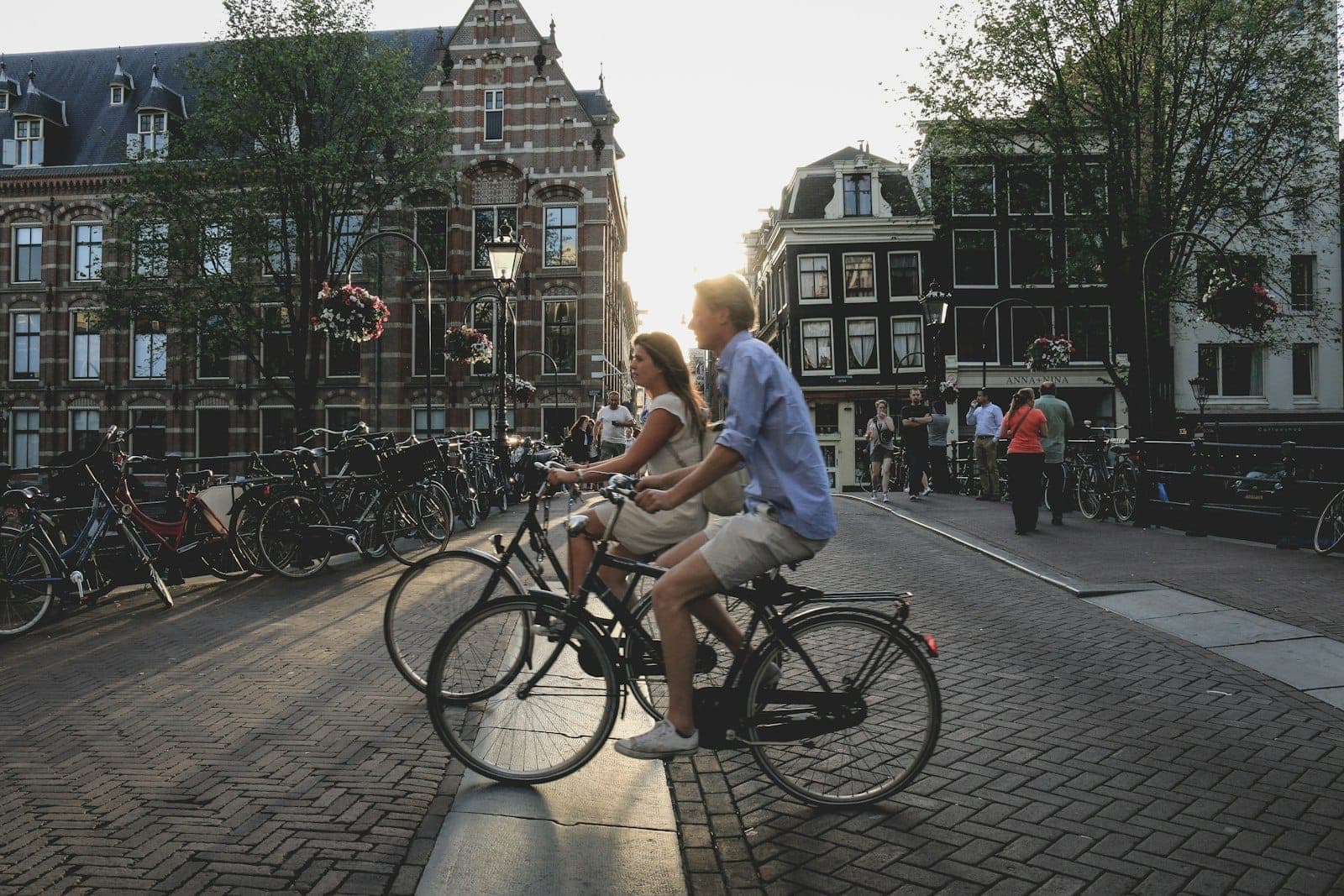 Ein Paar, das gemeinsam auf einer malerischen Straße in Amsterdam Fahrrad fährt, mit historischen Gebäuden und Fahrrädern bei Sonnenuntergang.