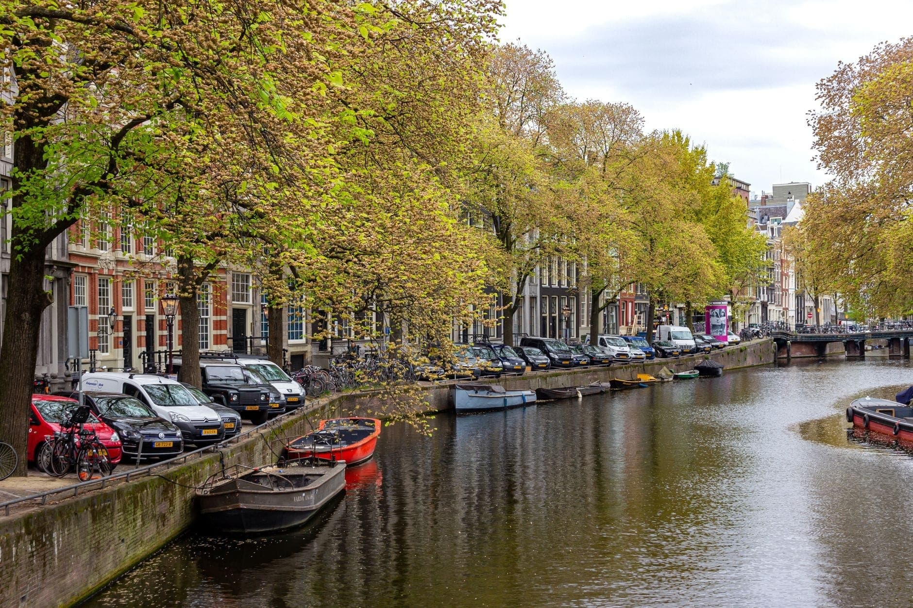 Baumgesäumter Amsterdamer Kanal mit vertäuten Booten und historischen Stadthäusern, der eine friedliche und romantische Nachbarschaftsszene für Paare zum Erkunden schafft.