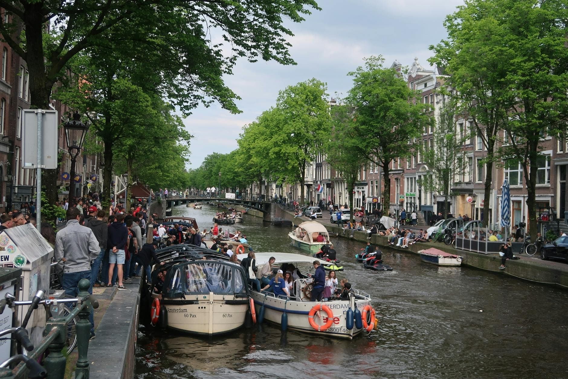 Amsterdamer Kanal voller Boote und Menschen an beiden Ufern, die Festivitäten genießen, lebhaft mit Gruppen und orangefarbenen Dekorationen.