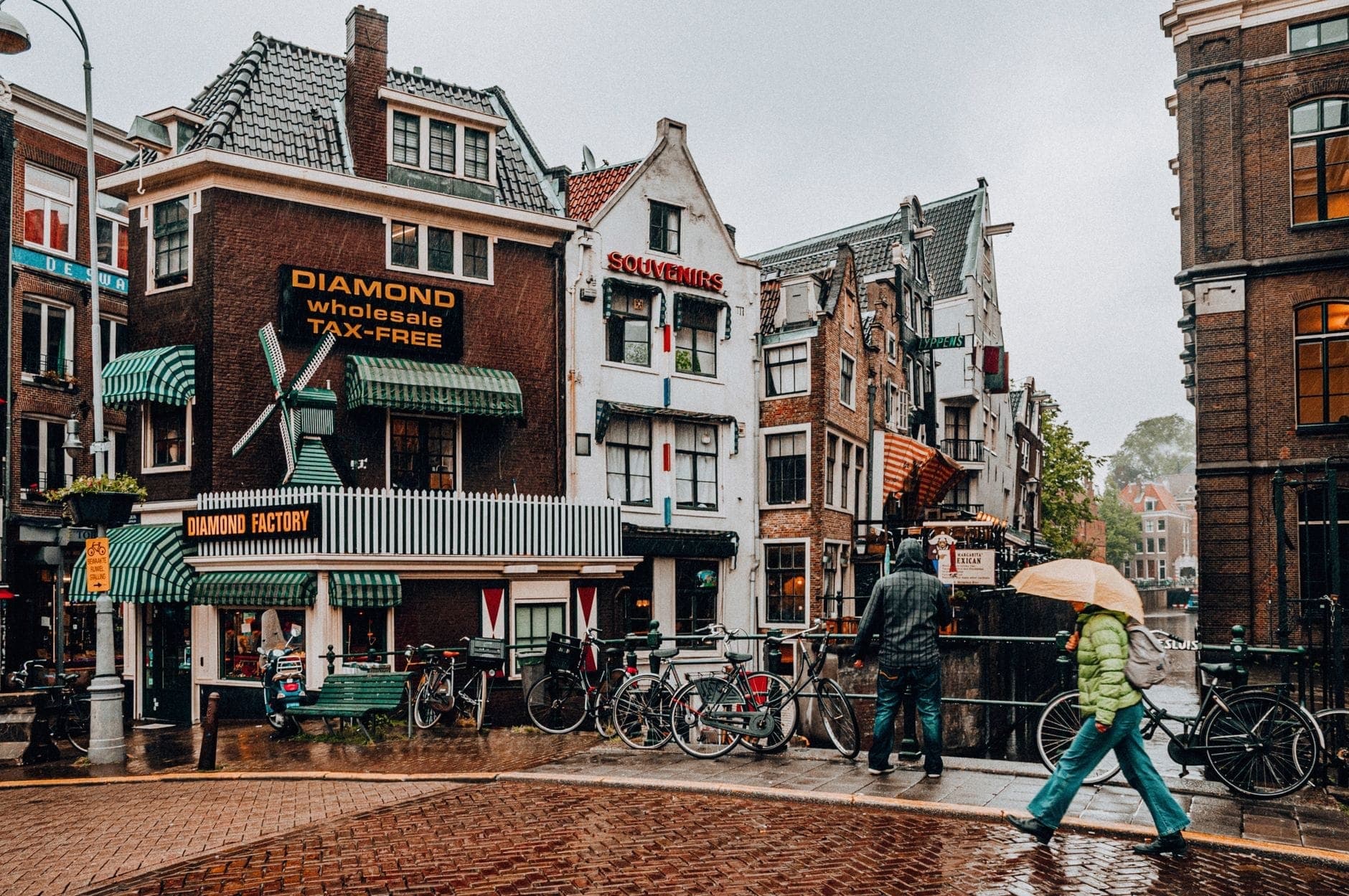 Menschen mit Regenschirmen bei Regenwetter auf einer Straße in Amsterdam mit traditionellen niederländischen Gebäuden im Hintergrund.
