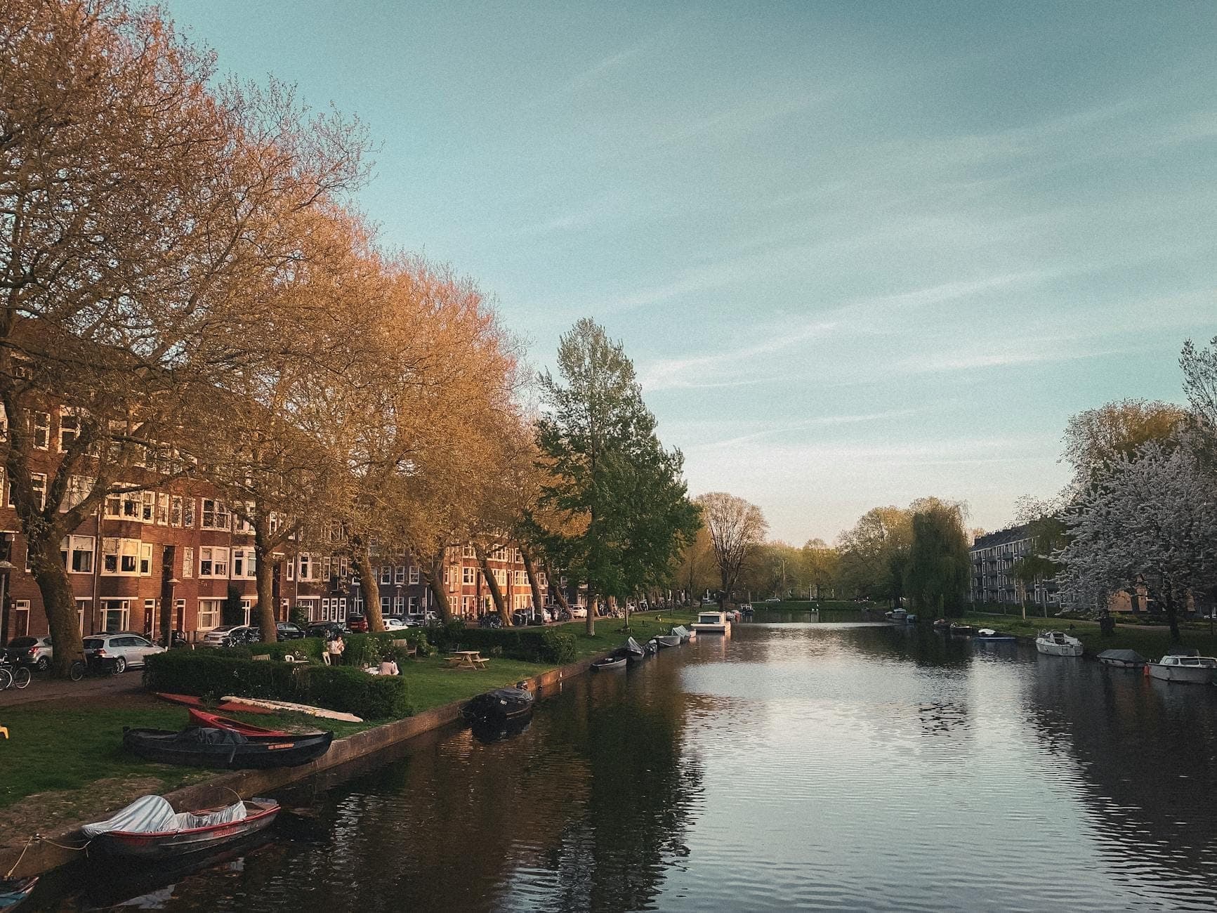 Ruhiger Amsterdamer Kanal im Frühling, gesäumt von blühenden Bäumen, vertäuten Booten und klassischen niederländischen Gebäuden unter blauem Himmel.