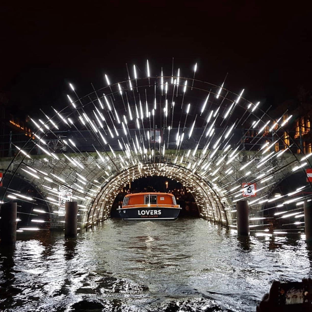 Beleuchtete Kunstinstallation beim Amsterdam Light Festival, mit einem Boot, das unter einer mit strahlend weißen Lichtern beleuchteten Brücke über den Kanal bei Nacht hindurchfährt.
