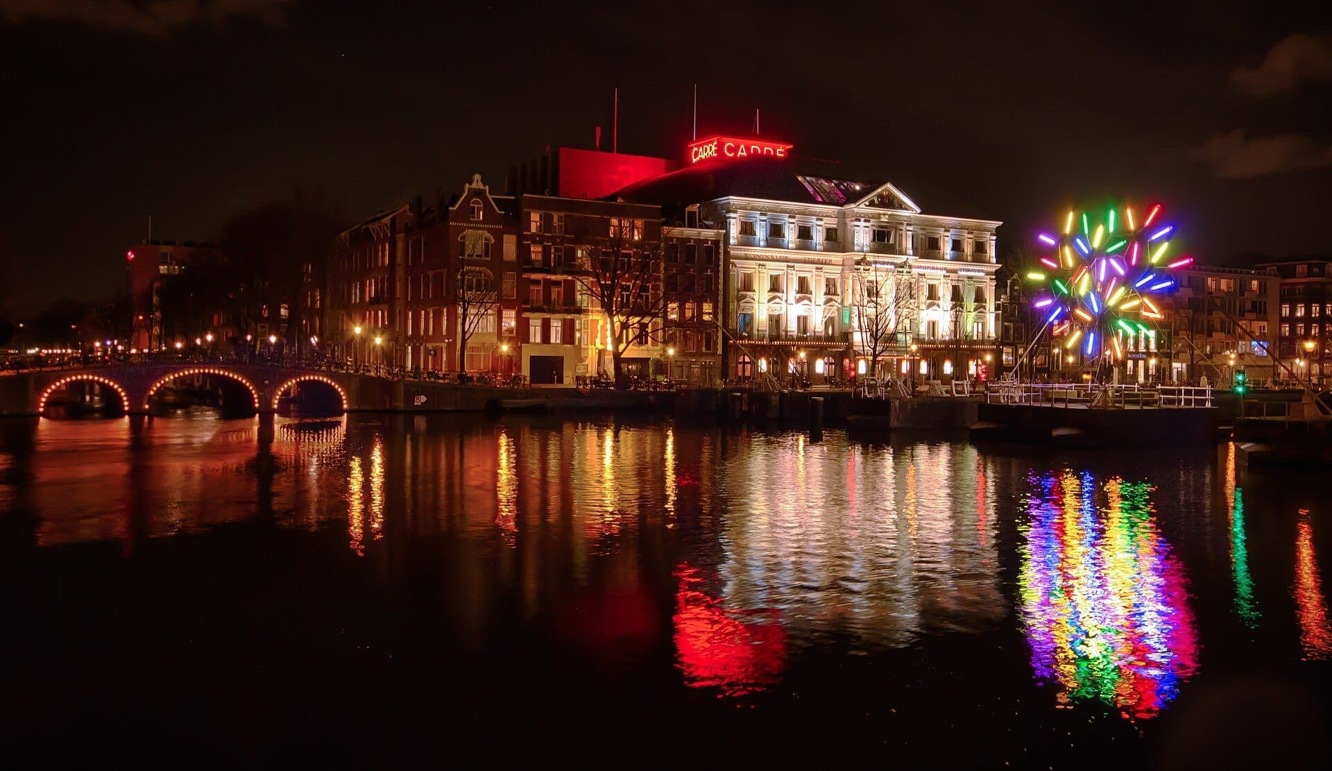 Beleuchtete historische Gebäude und Kanalbrücke spiegeln sich im Wasser während des Amsterdam Light Festival wider, mit einer farbenfrohen Lichtinstallation am Ufer.