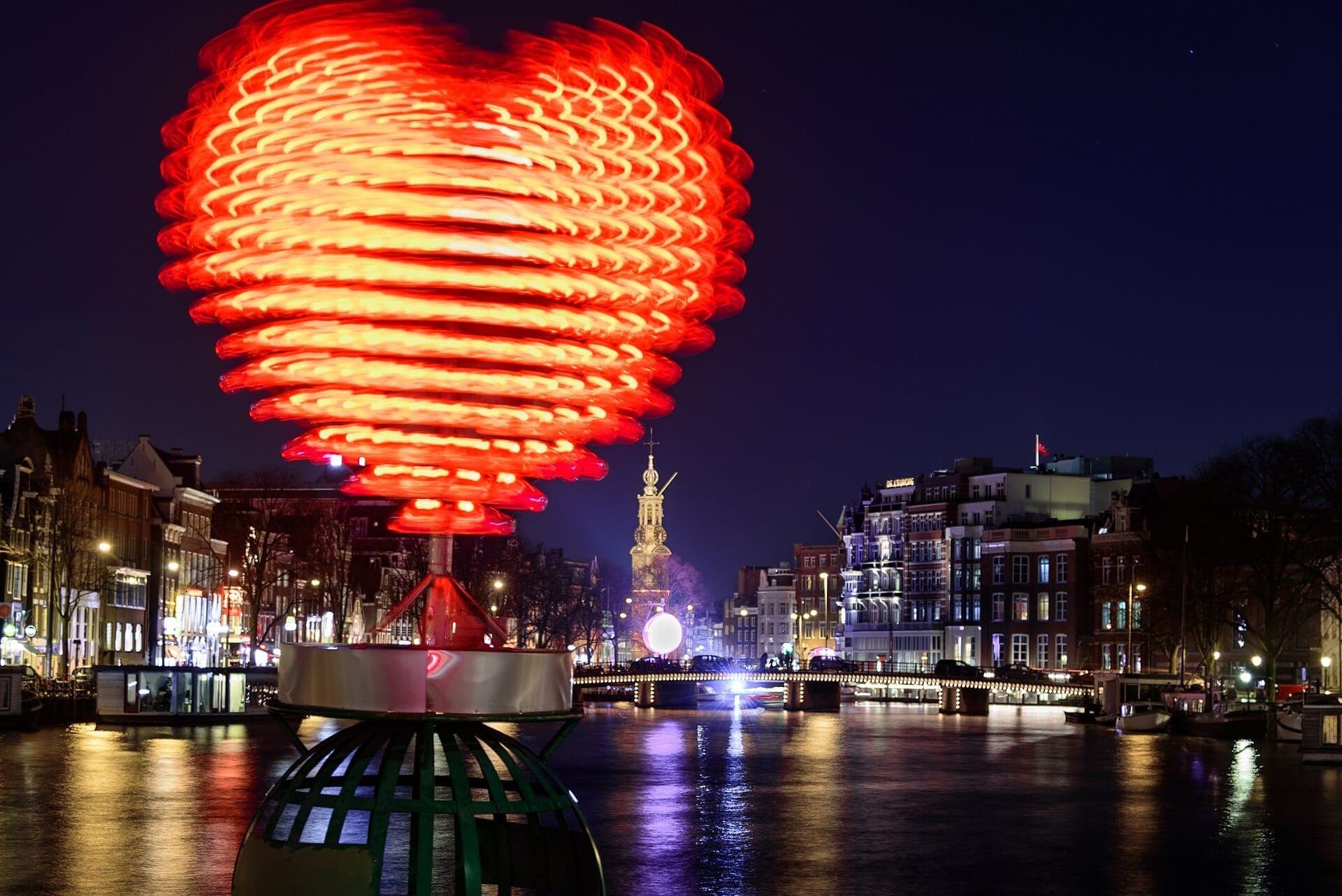 Eine große beleuchtete herzförmige Lichtskulptur leuchtet nachts über einem Amsterdamer Kanal beim Amsterdam Light Festival, mit historischen Gebäuden und Spiegelungen im Wasser.