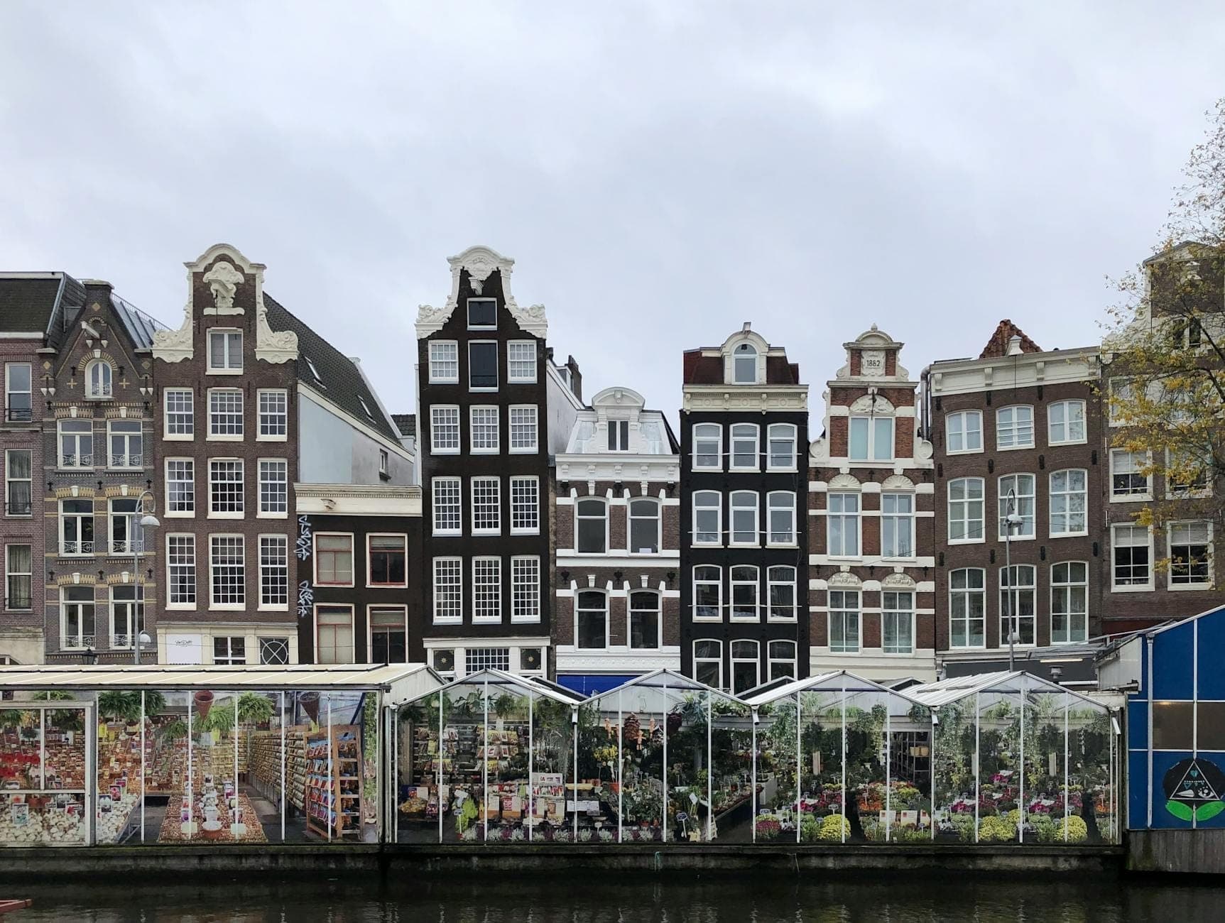 Blick auf die ikonischen Grachtenhäuser von Amsterdam hinter den berühmten schwimmenden Blumenständen des Bloemenmarkt am Singel-Kanal unter bewölktem Himmel.