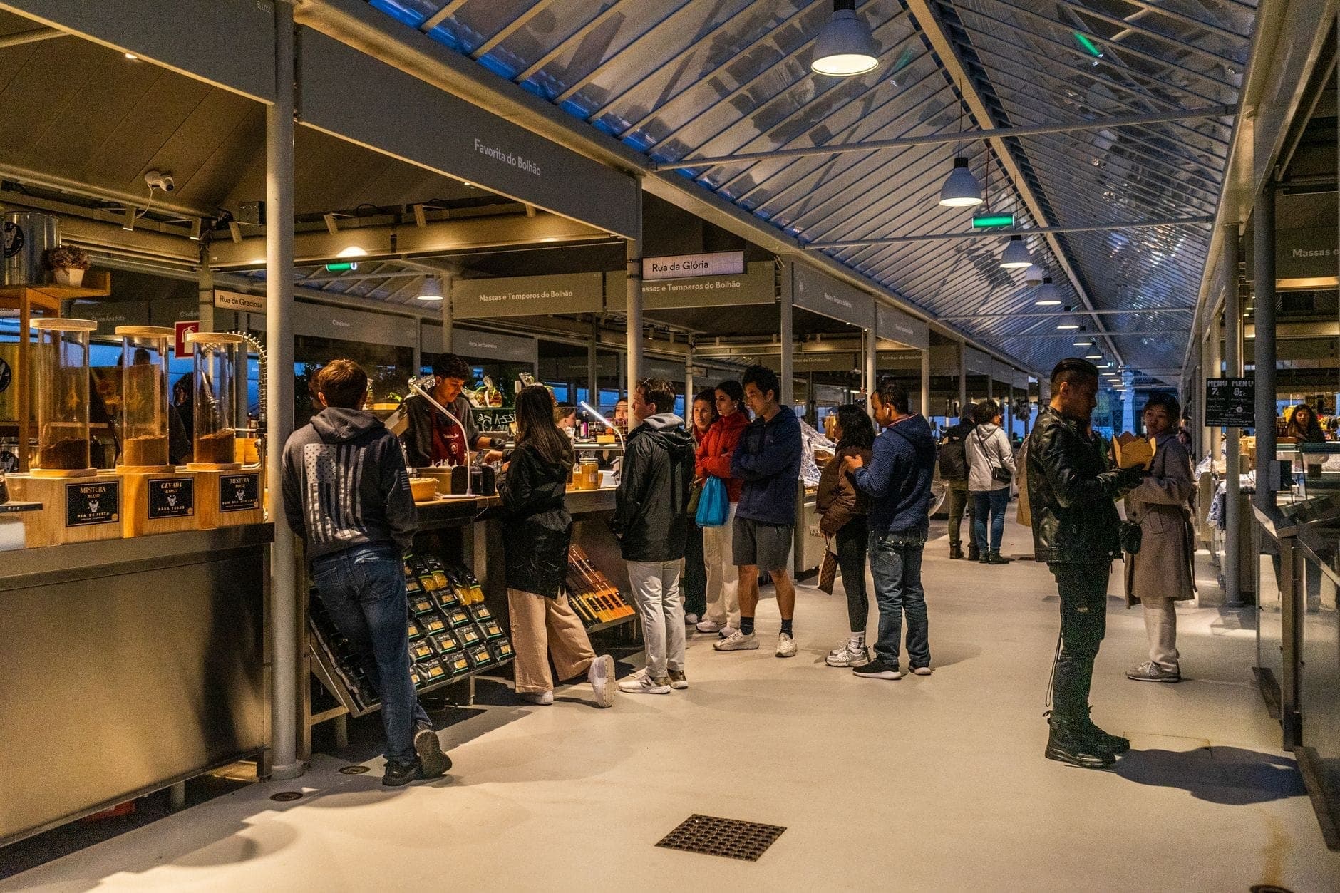 Menschen stehen und bestellen Essen an Marktständen in einer überdachten Markthalle unter einem modernen Glasdach in Amsterdam.