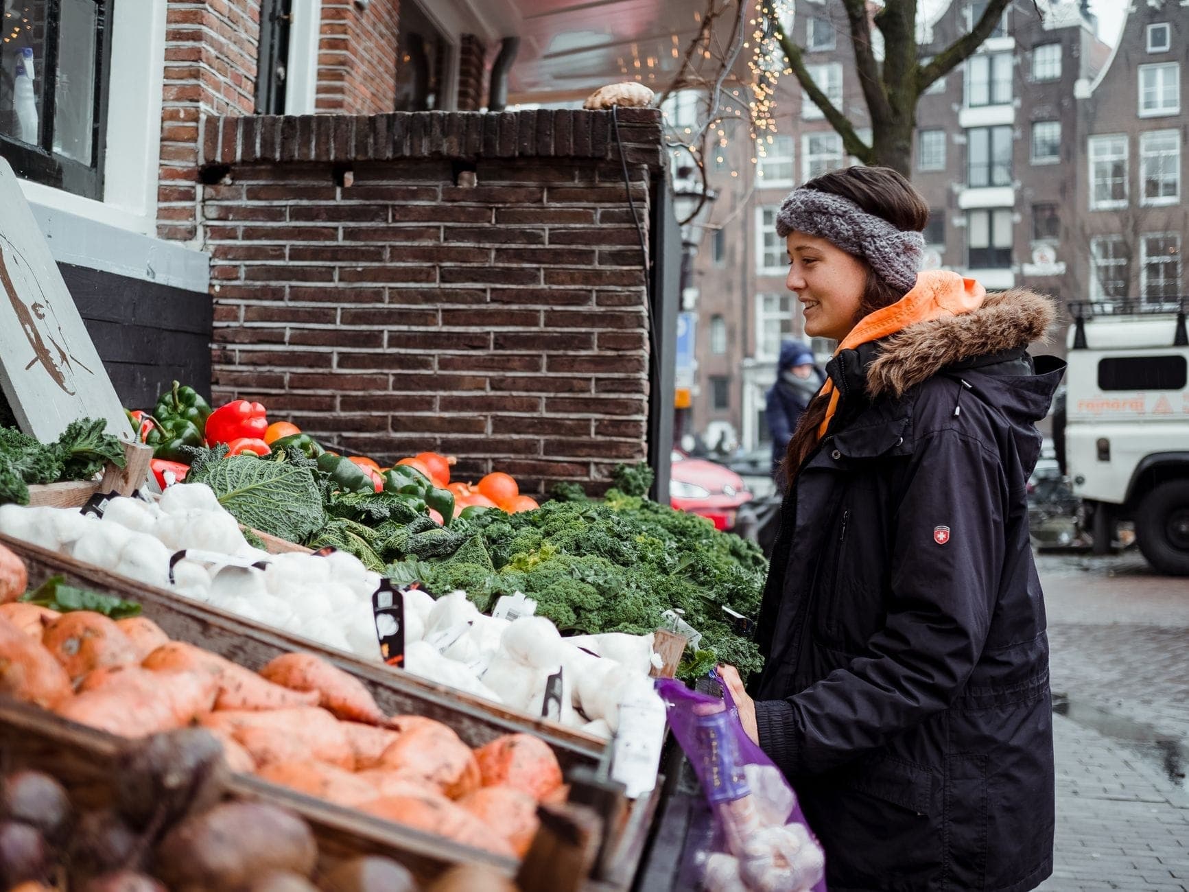 Junge Frau, die lächelnd an einem Gemüsestand auf einem Stadtmarkt einkauft, mit Amsterdam-typischen Backsteingebäuden im Hintergrund.