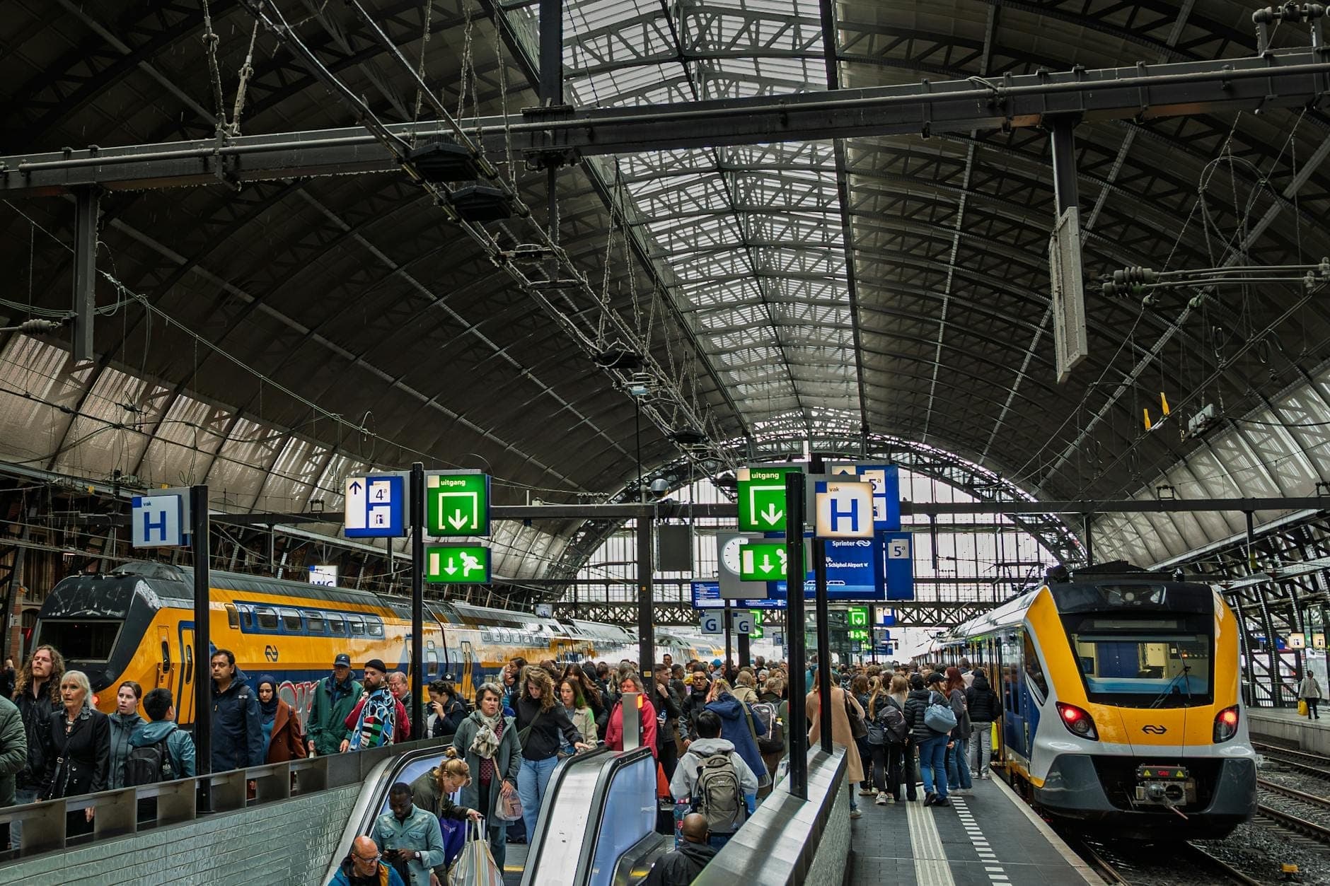Überfüllte Bahnsteige und Züge im Inneren des Bahnhofs Amsterdam Centraal unter einem großen gewölbten Glasdach, mit übersichtlicher Beschilderung und Rolltreppen.