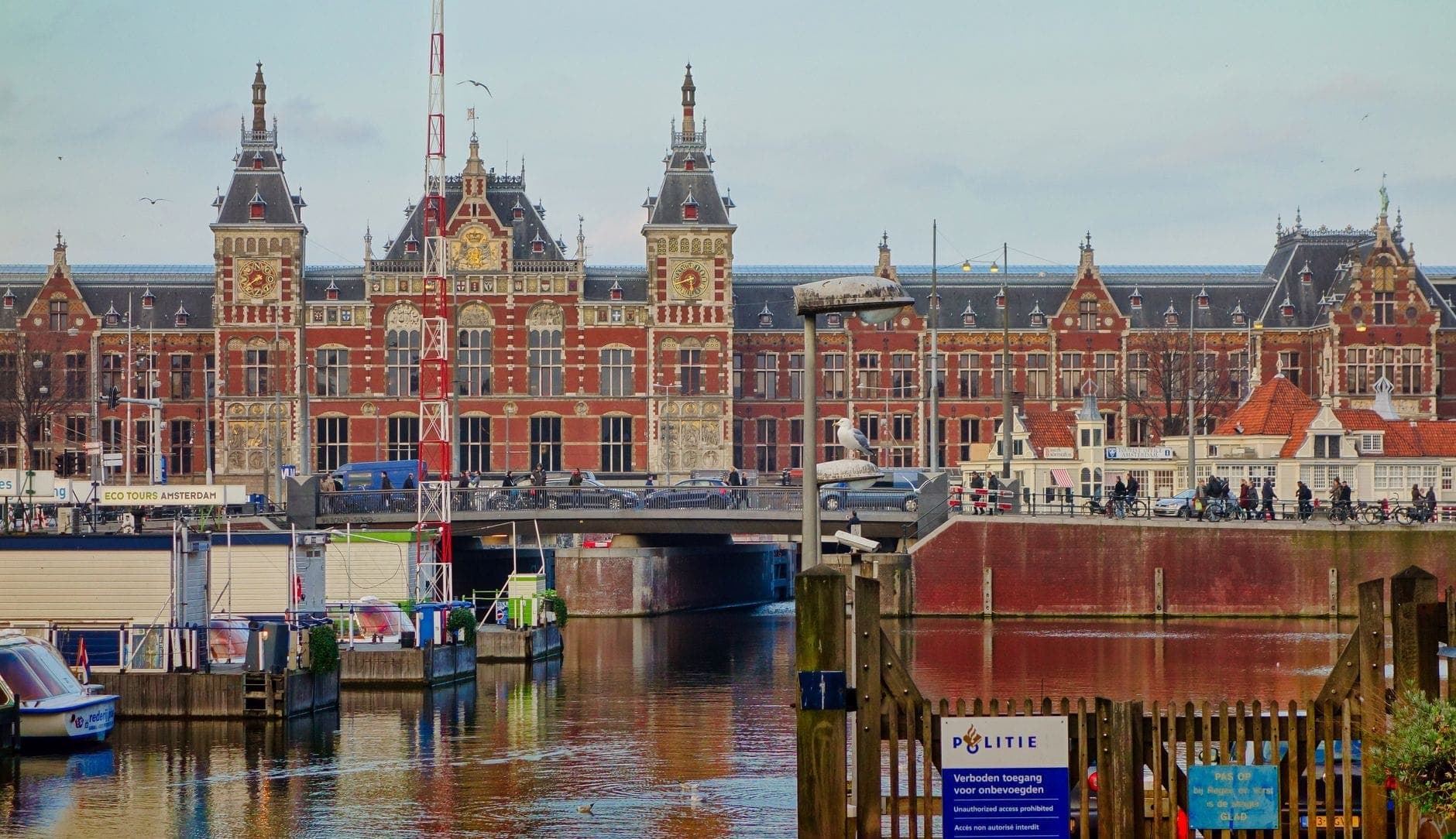 Außenansicht des Bahnhofs Amsterdam Centraal mit verzierter Architektur, Stadtkanal im Vordergrund und Personen am Eingang.
