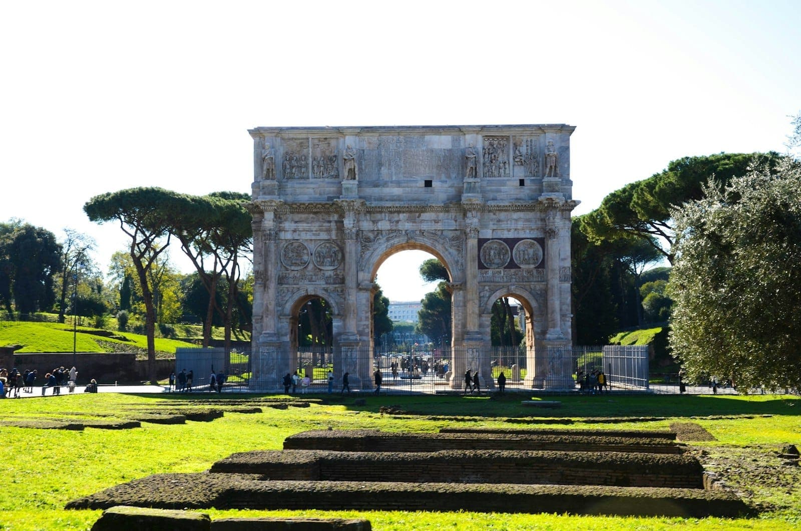 L'Arco di Costantino a Roma, un imponente arco di trionfo romano circondato da prato verde brillante e rovine in pietra, sotto un cielo terso e soleggiato.