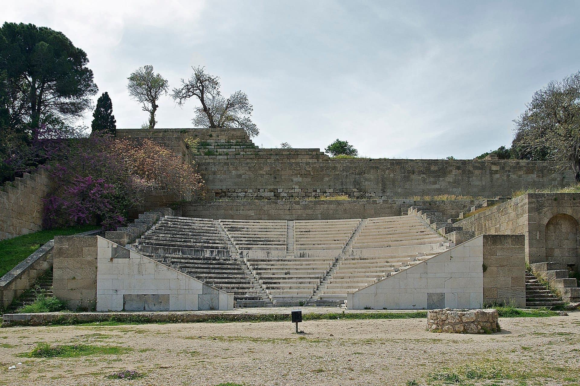 Sièges en pierre restaurés de l'Ancien Stade de Rhodes avec des marches usées, entourés de vieux murs en pierre et d'arbres épars sous un ciel nuageux.