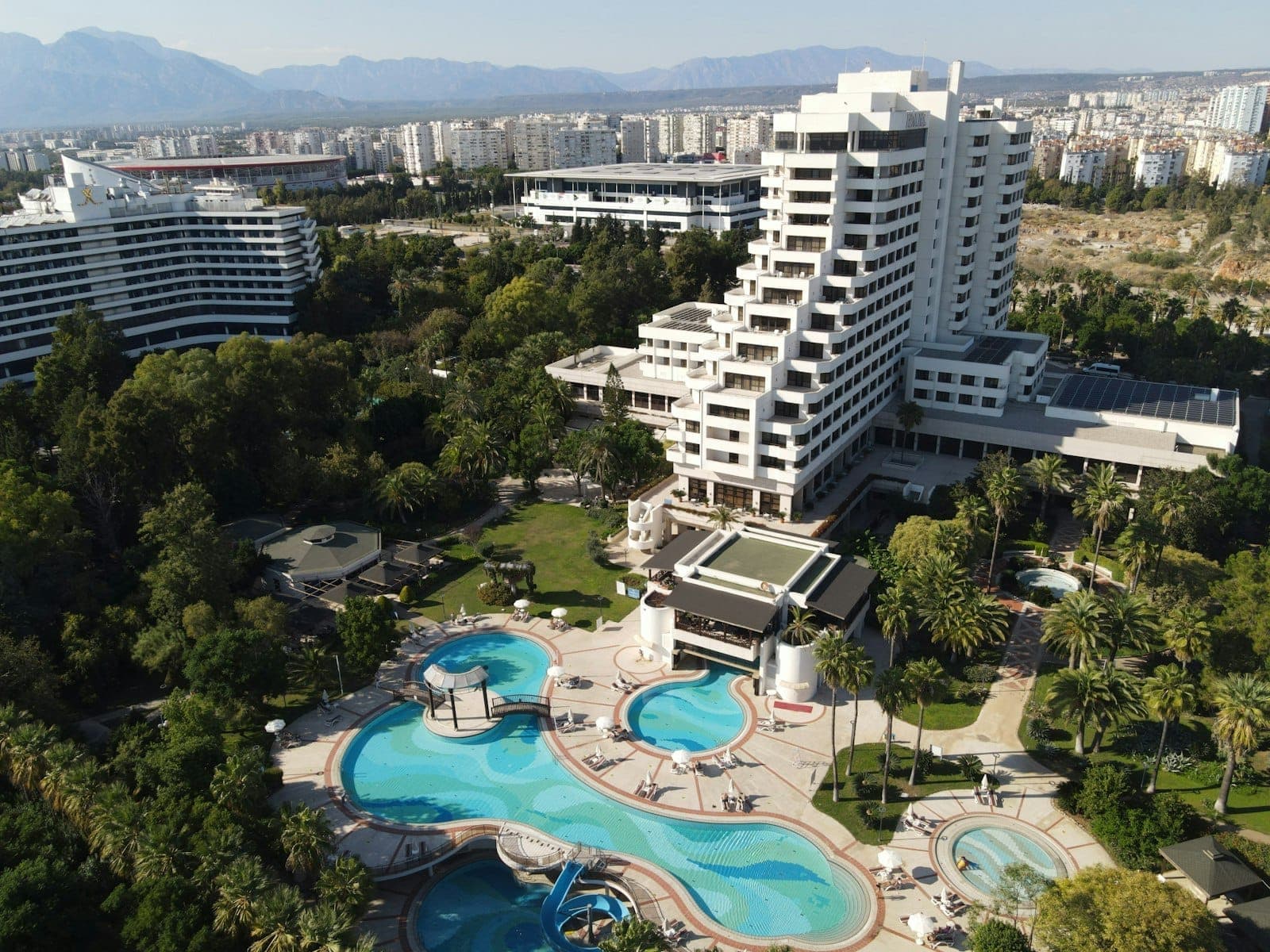 Aerial view of a large Antalya resort hotel with pools, gardens, and cityscape, mountains visible in the distance.