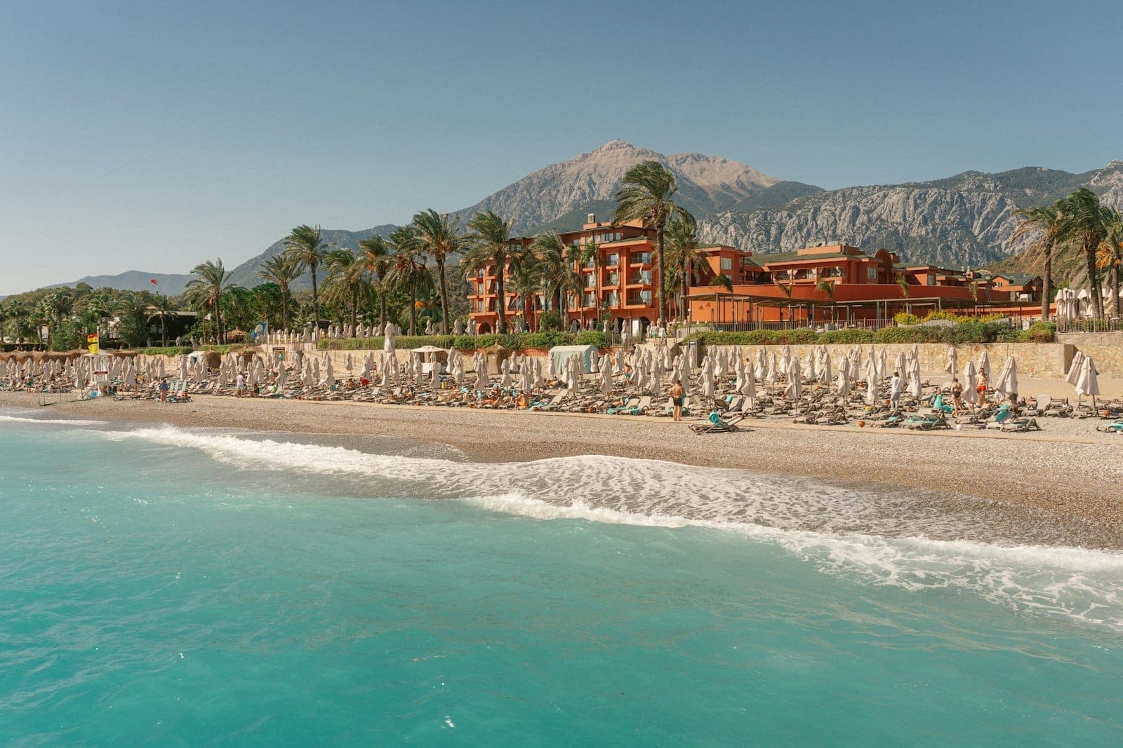 Ein Strandresort in Antalya mit Reihen von Liegestühlen, Palmen, einem großen Hotelgebäude und Bergen im Hintergrund unter klarem Himmel.