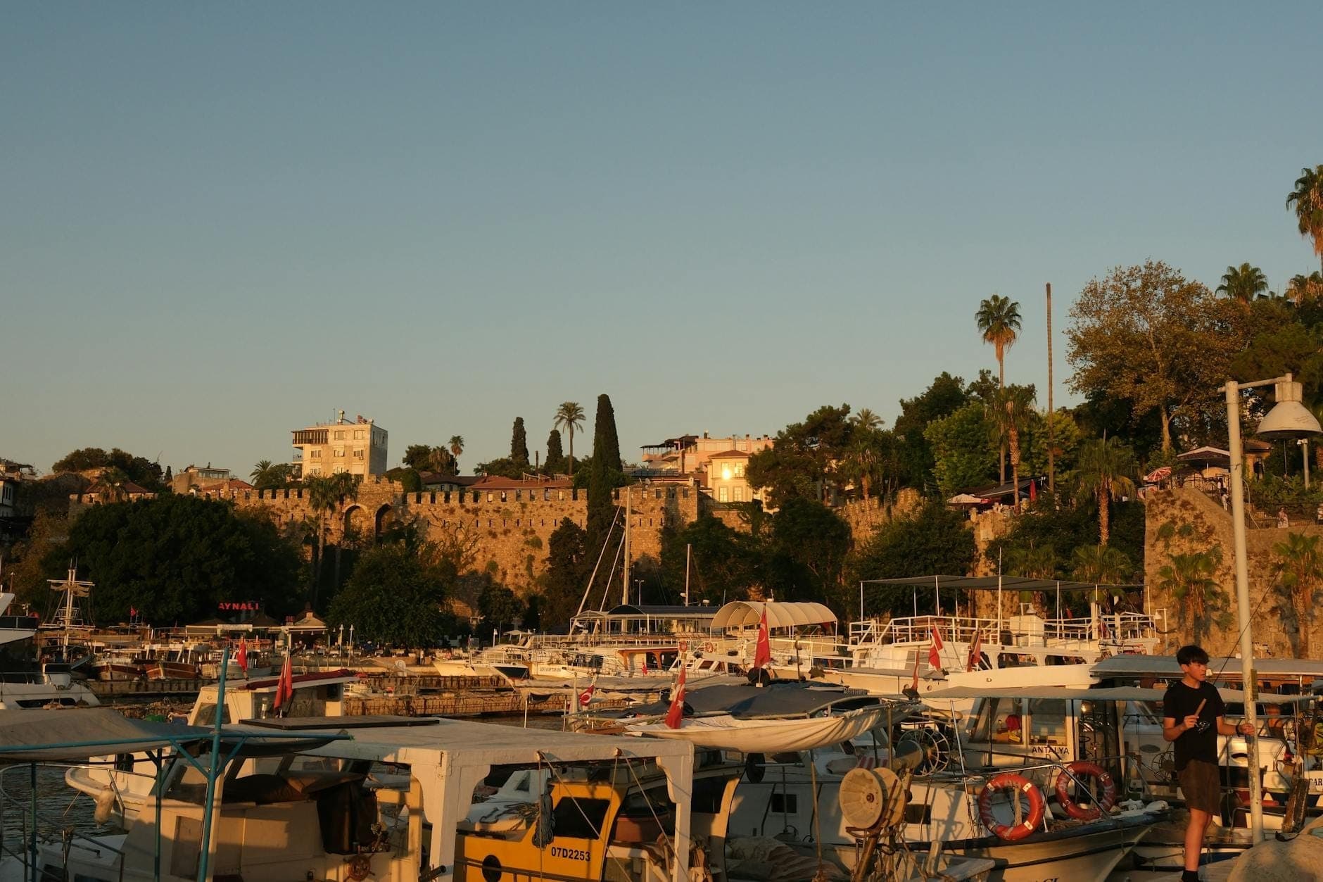 Antalya harbor at sunset with boats docked and historic city walls and greenery in the background, capturing the area’s coastal charm.